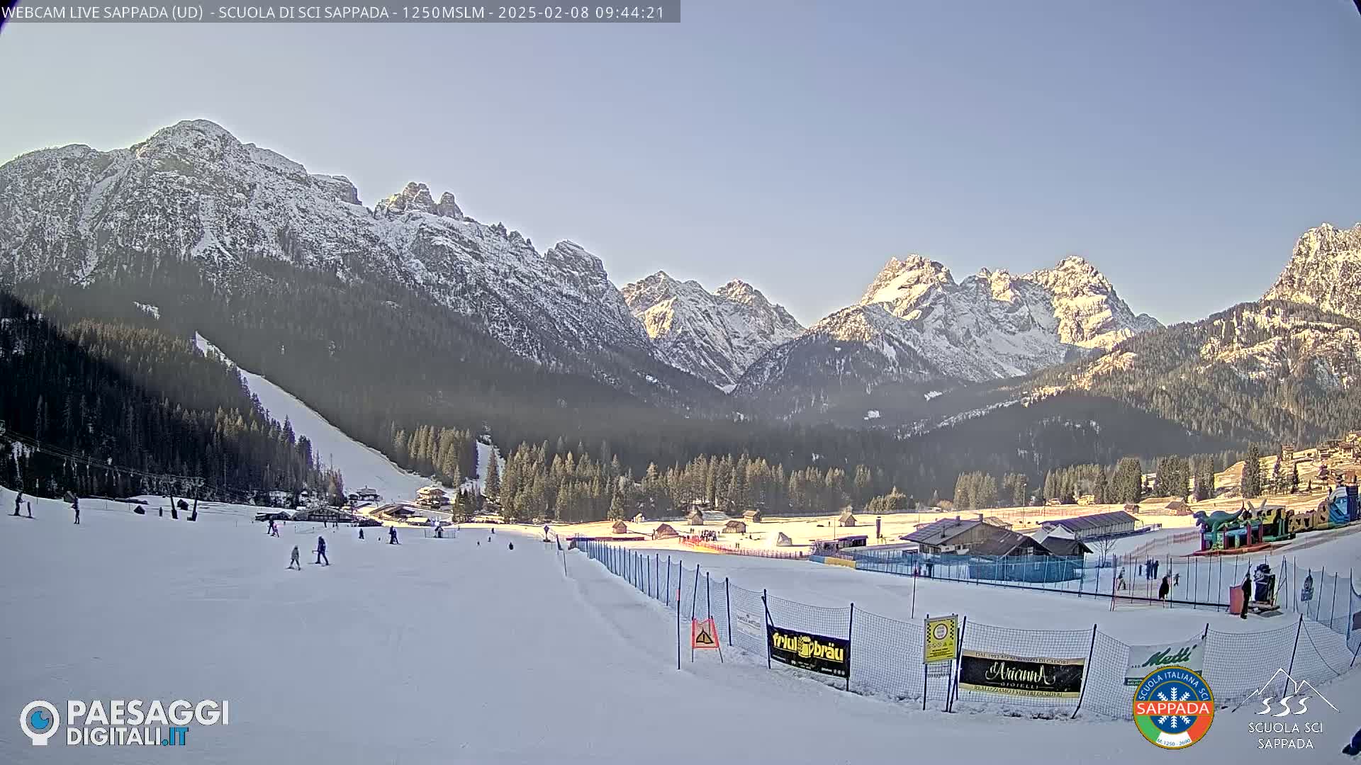 A snow-covered ski slope with a few skiers, nestled in a valley surrounded by snow-capped mountains under a clear blue sky.