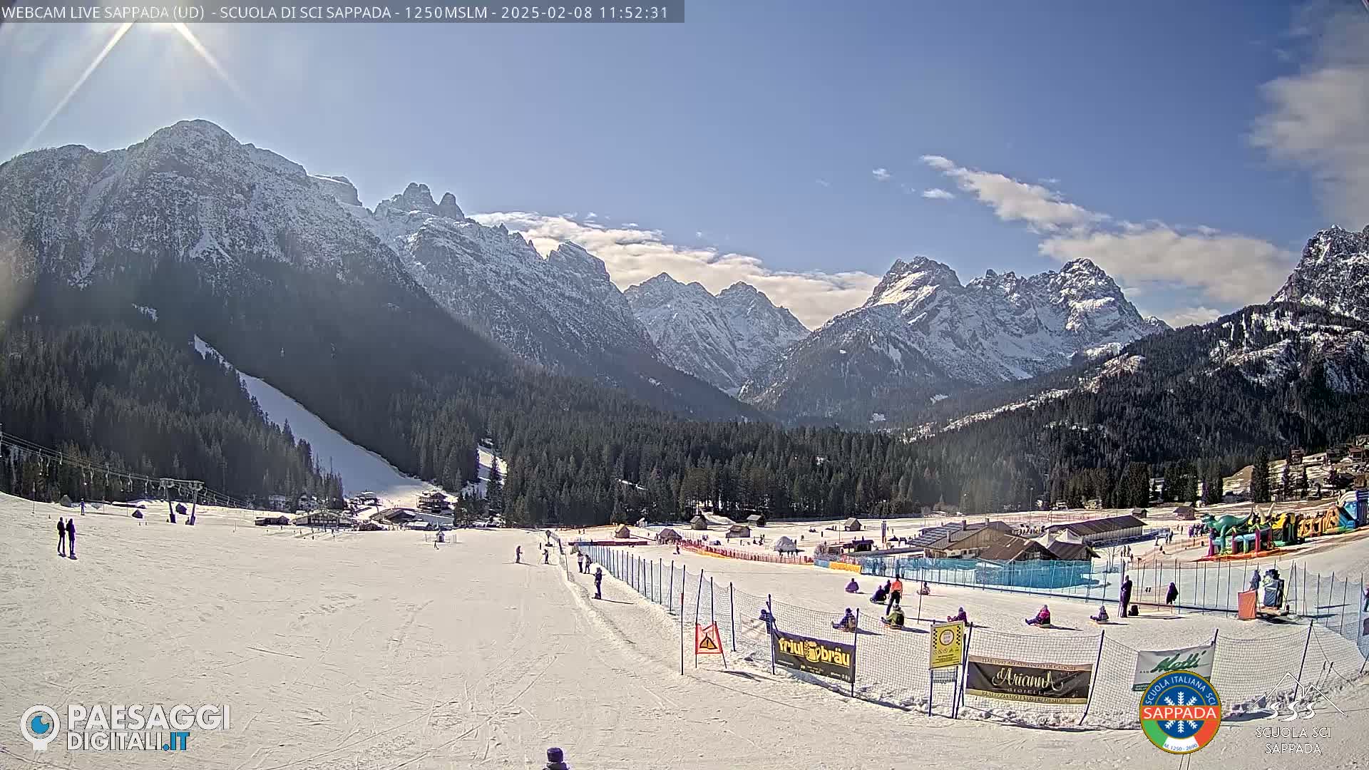 A snow-covered ski slope with people skiing and playing, situated in a valley surrounded by snow-capped mountains under a mostly sunny sky.