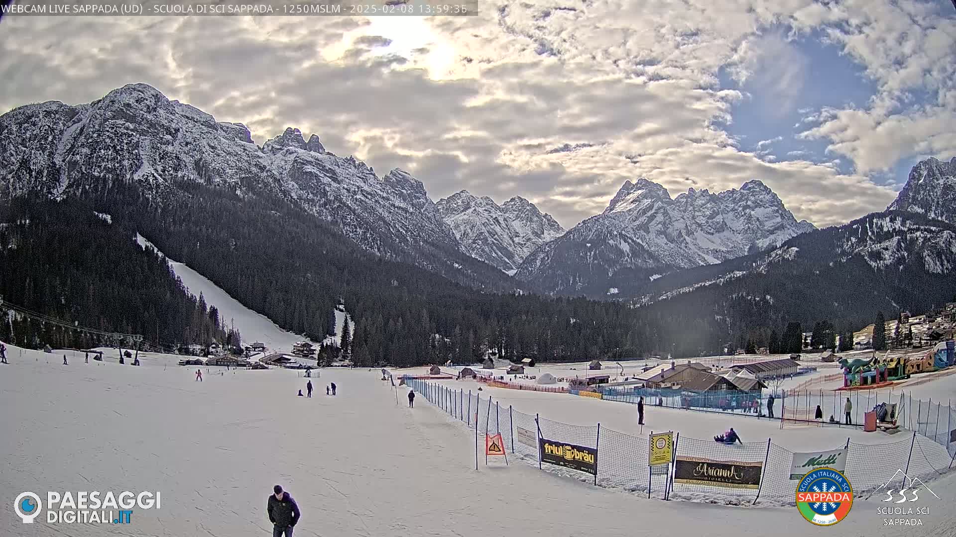 A snow-covered ski slope with a few people on it sits in a valley between snow-capped mountains under a partly cloudy sky.