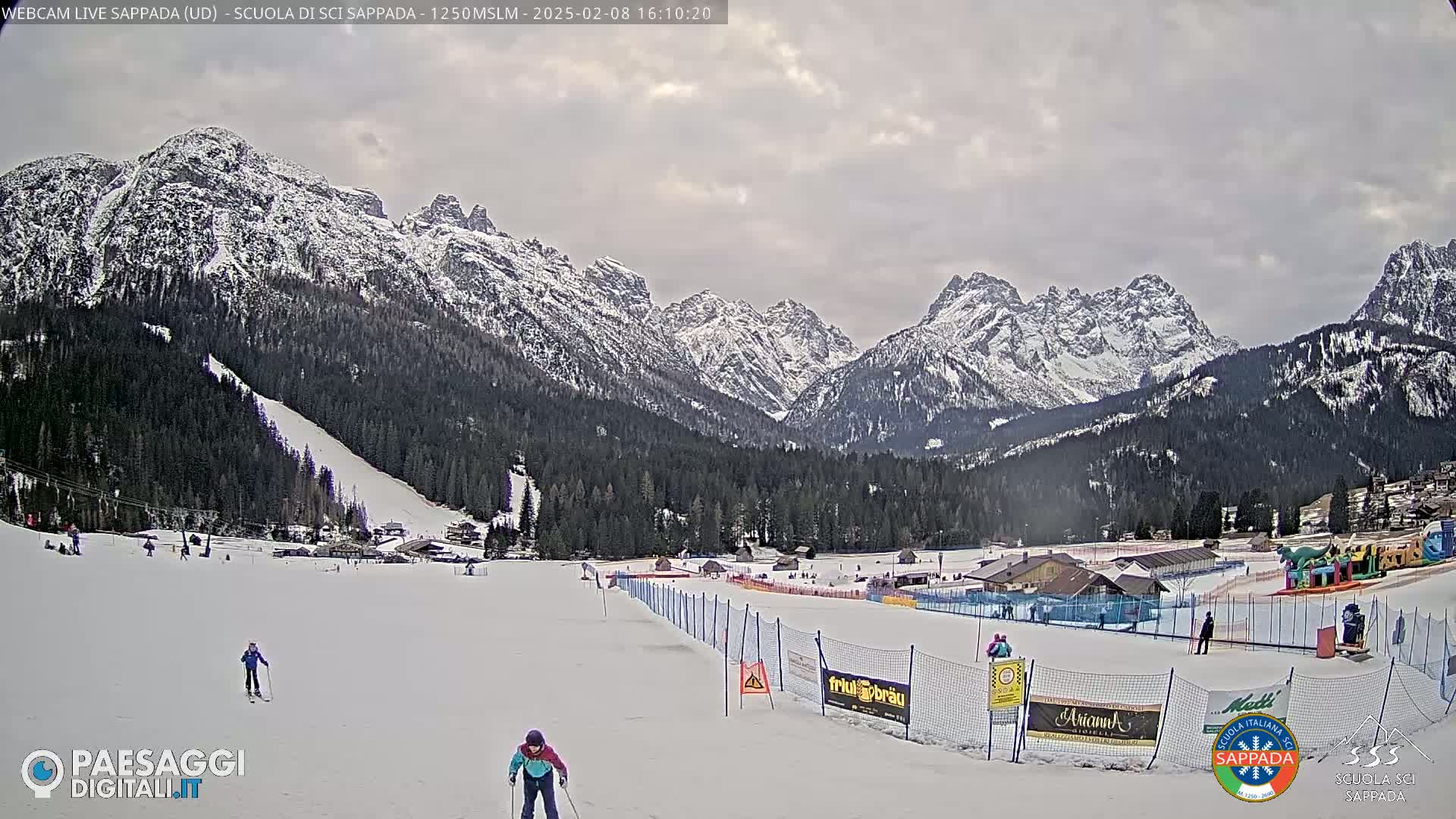 A snowy ski slope with skiers and a children's play area is nestled in a valley surrounded by snow-capped mountains under an overcast sky.