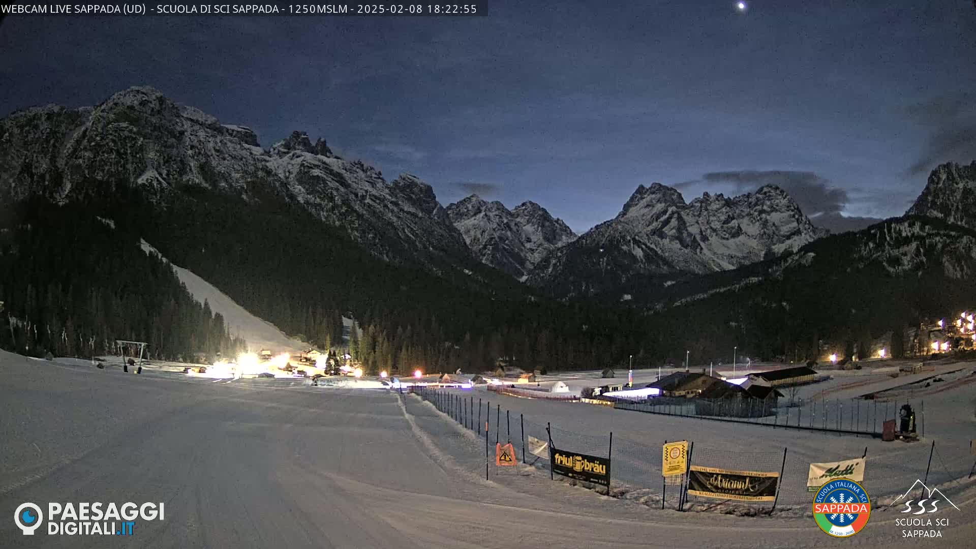 A snow-covered ski resort at night, illuminated by lights, is nestled in a valley surrounded by snow-capped mountains under a twilight sky.