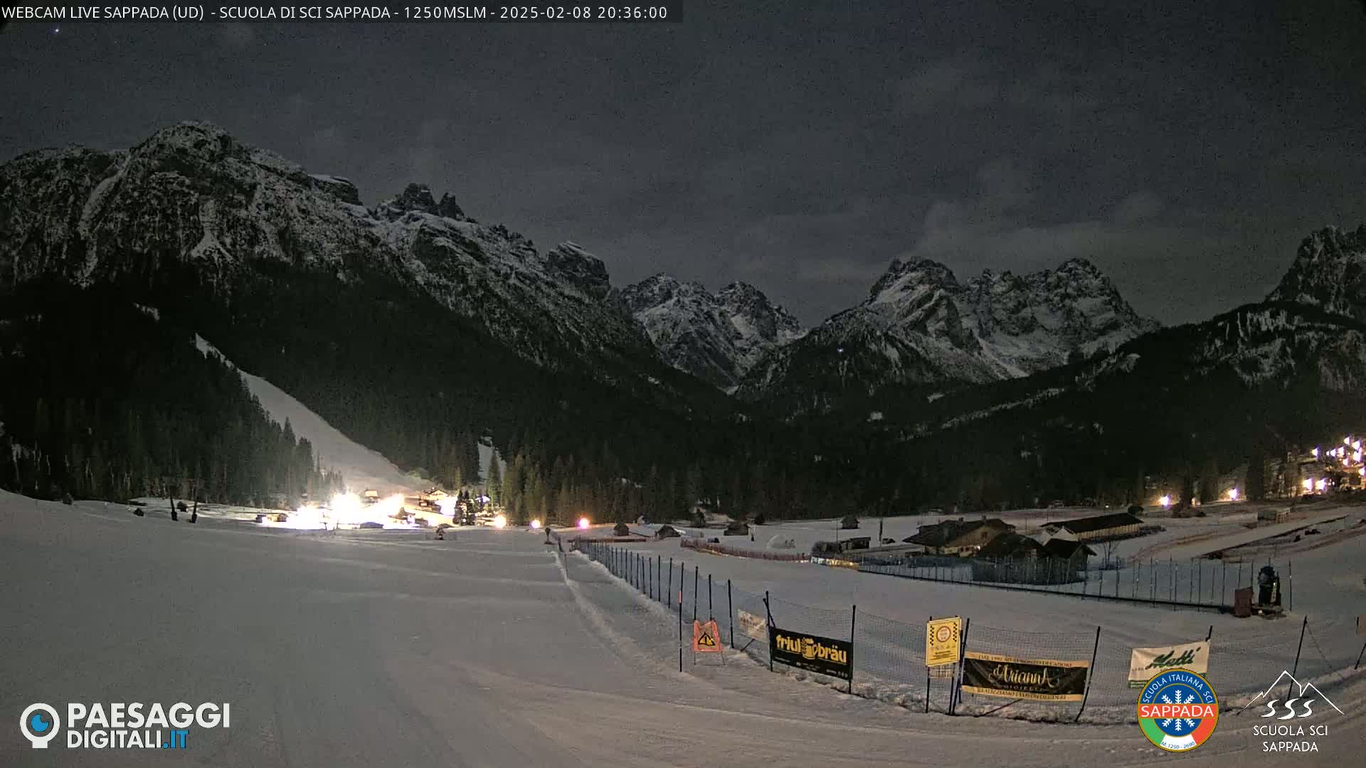 A snow-covered ski slope at night, illuminated by lights, is nestled in a valley between snow-capped mountains under a dark sky.