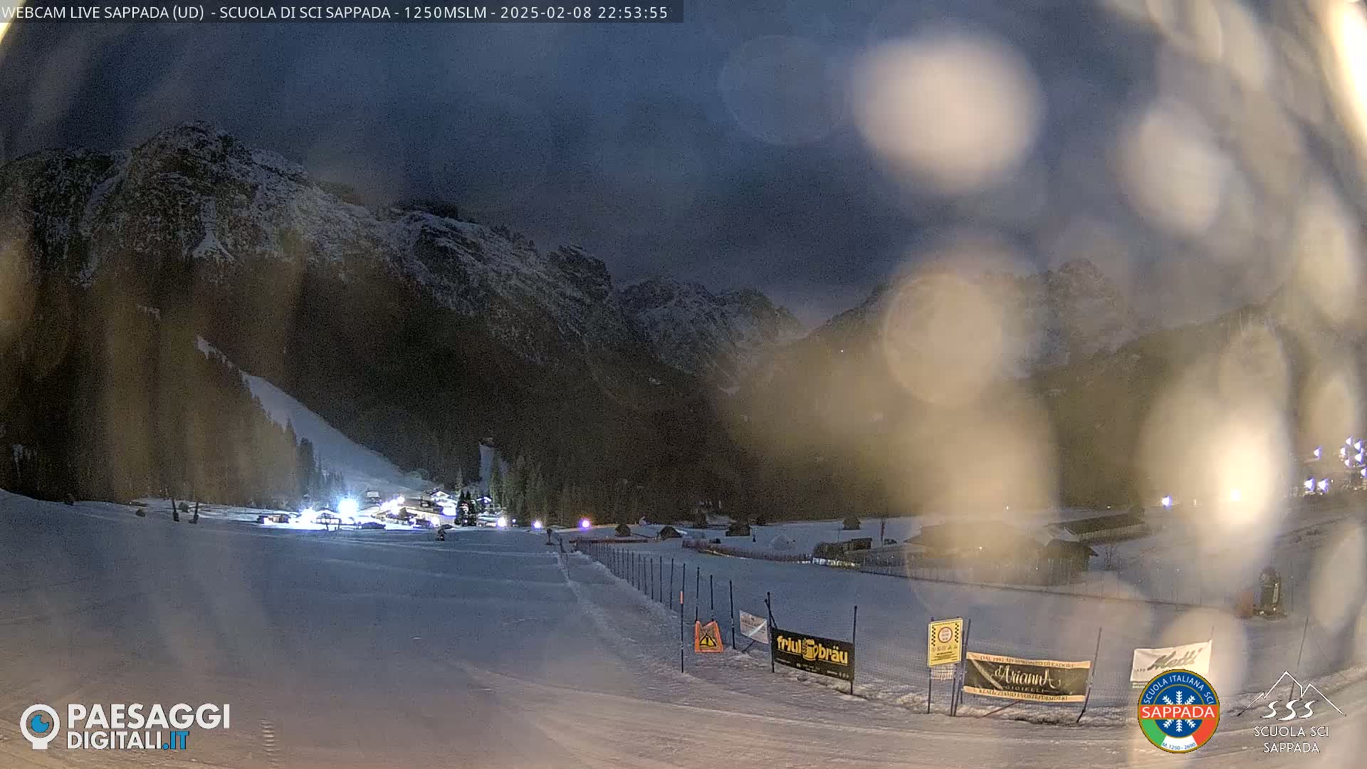 A nighttime view of a snow-covered ski slope at the base of snow-capped mountains under a dark, possibly overcast sky.