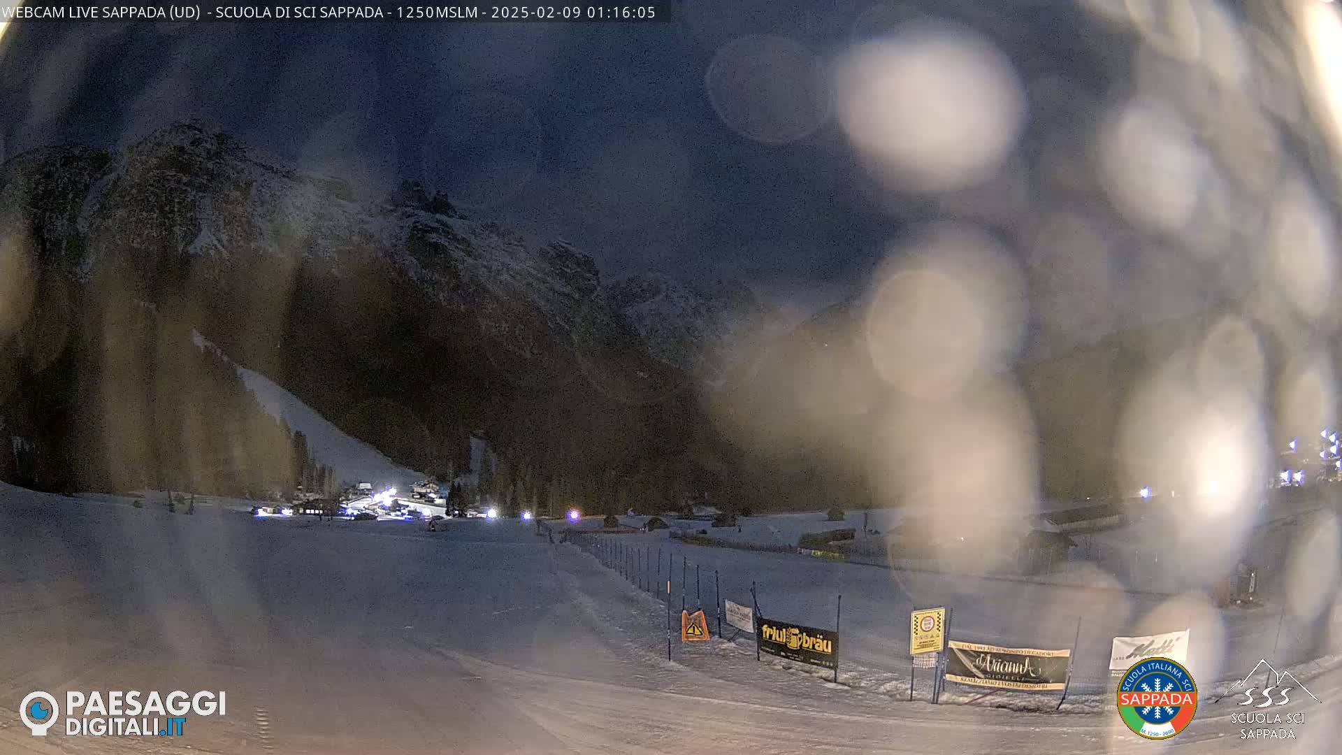 A nighttime view of a snow-covered ski slope with a mountain in the background under a dark, possibly cloudy sky.