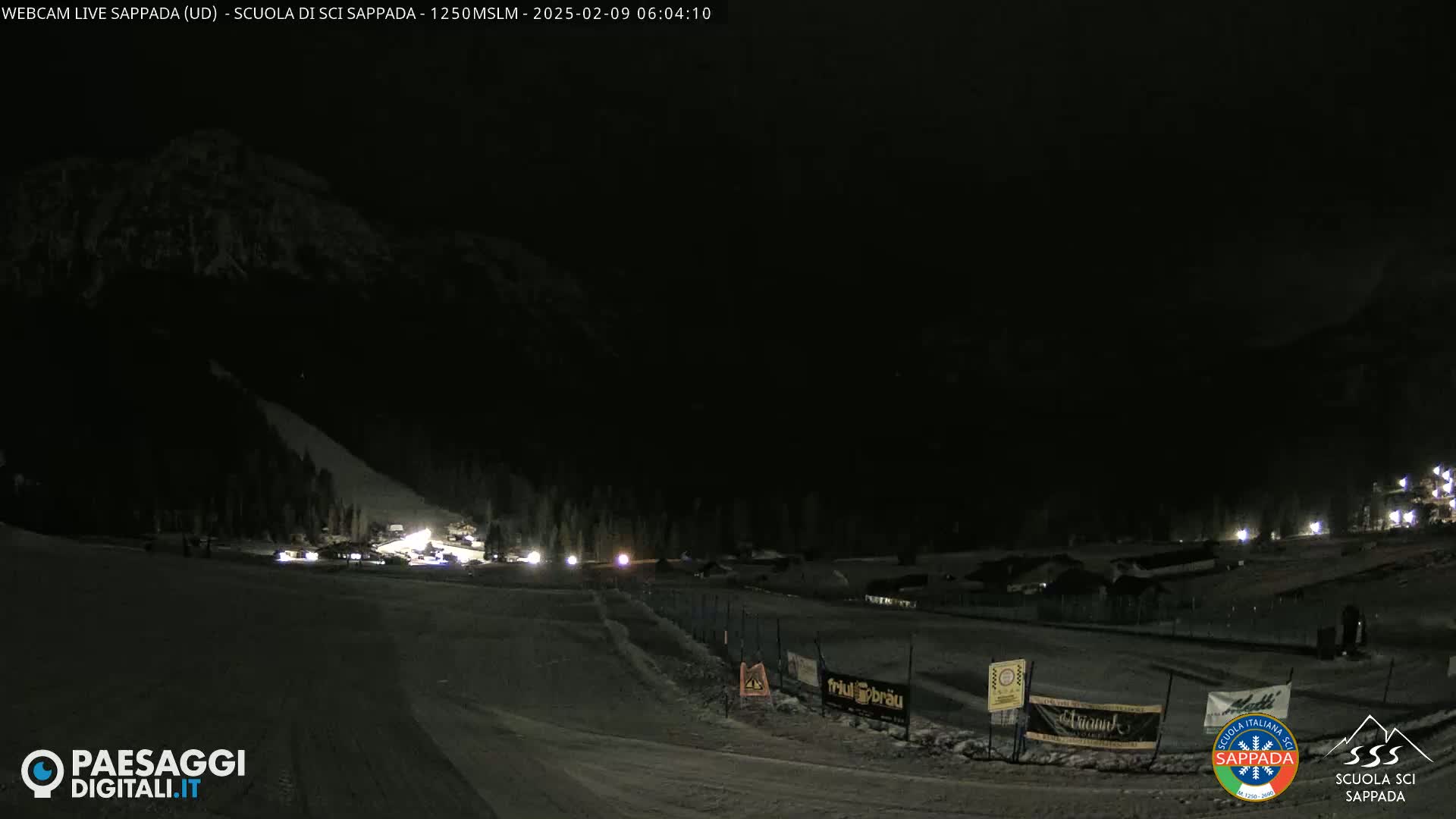 A nighttime view of a snow-covered ski slope with dimly lit buildings and vehicles in the distance, under a dark sky.
