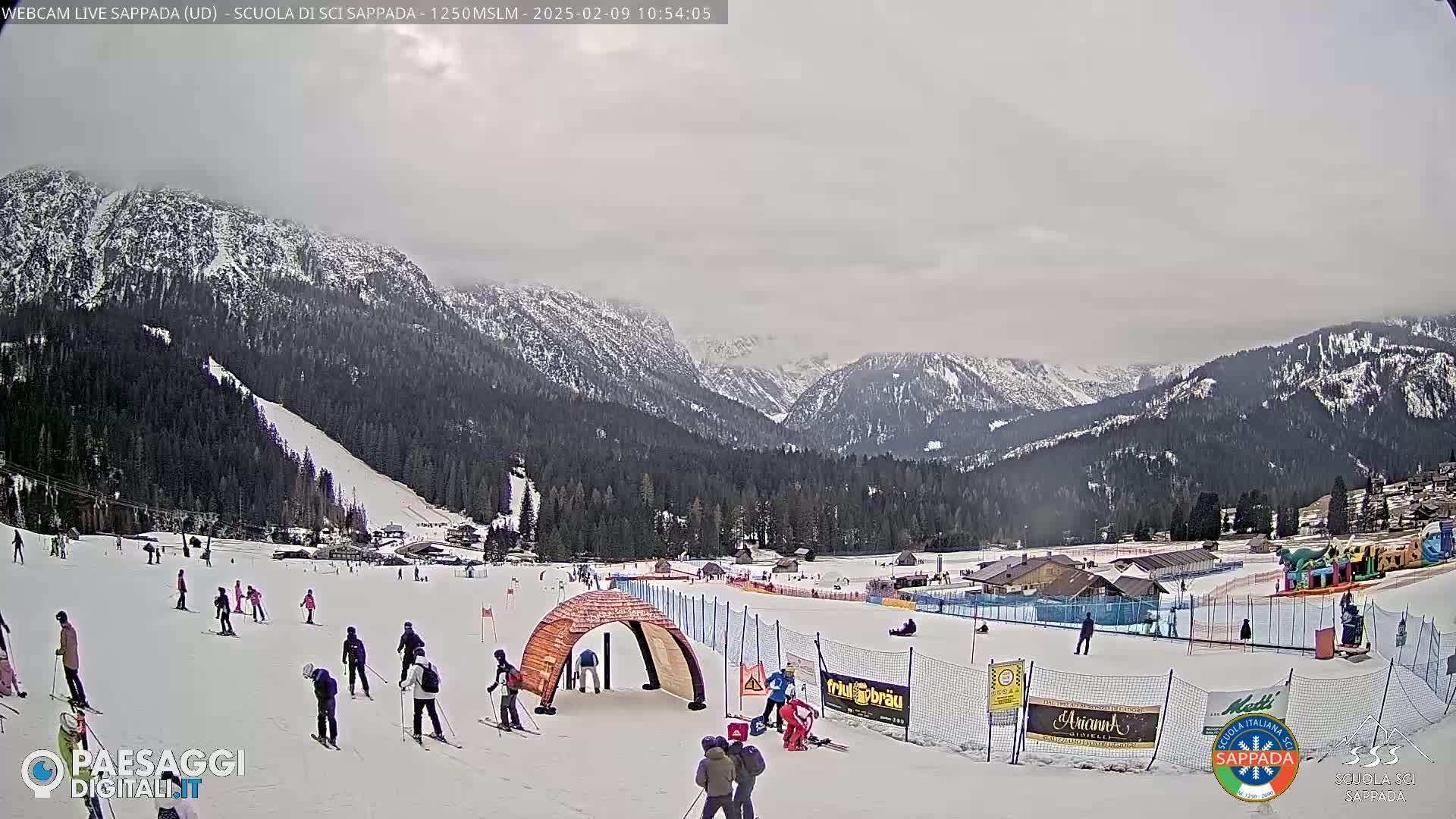 A snowy ski slope with skiers, a wooden archway, and mountain scenery under an overcast sky.