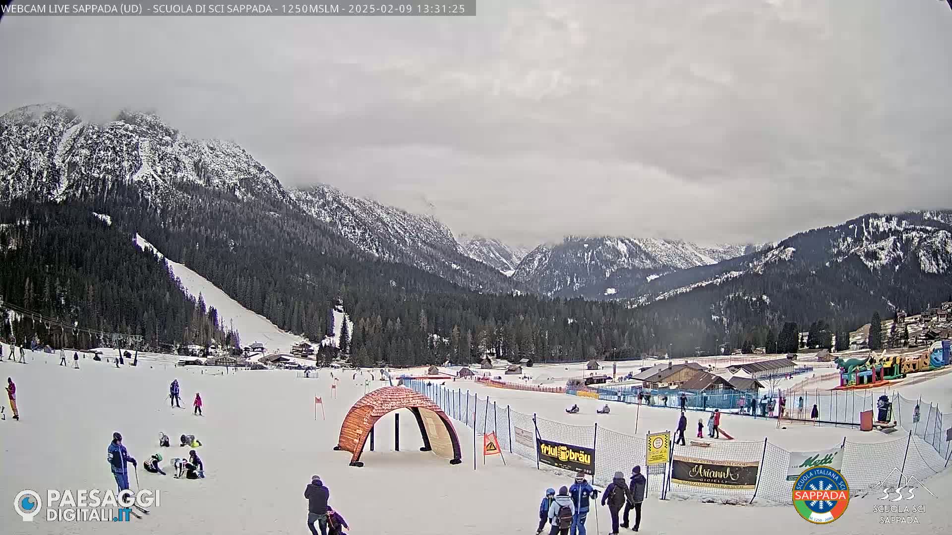 A snowy ski slope with people skiing and playing, situated in a valley surrounded by snow-covered mountains under an overcast sky.