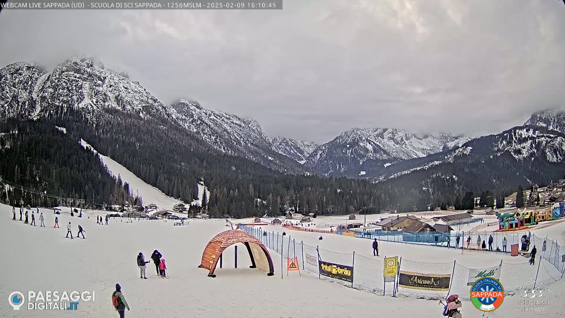 A snowy mountain ski resort scene under an overcast sky shows skiers and snowboarders on a slope with buildings and trees in the background.