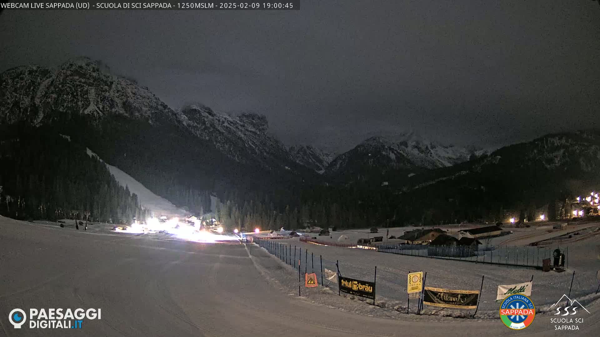 A snow-covered ski slope at night is illuminated under a dark, overcast sky, with mountains in the background.