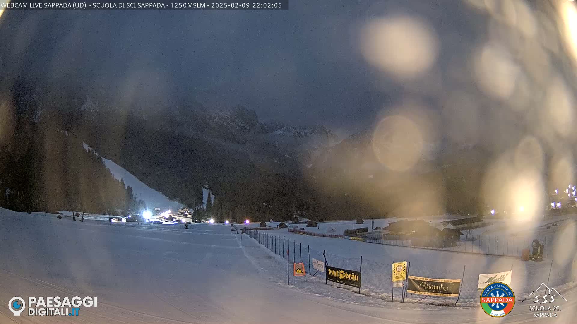 A snow-covered ski slope at night, with mountains in the background under a dark, hazy sky.
