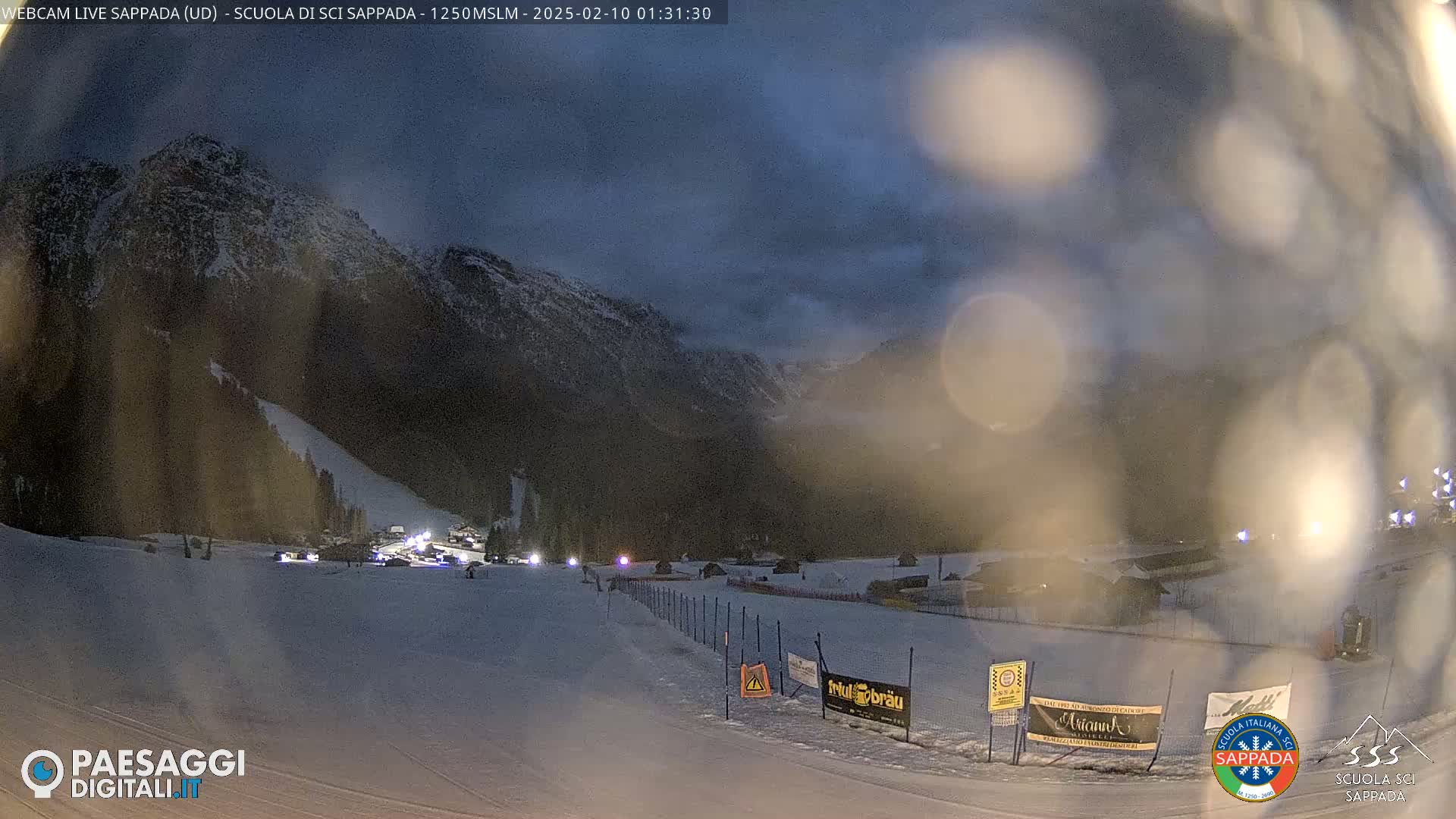 A snow-covered ski slope at night under a dark, cloudy sky, with mountains in the background and lights illuminating a lower section of the slope.