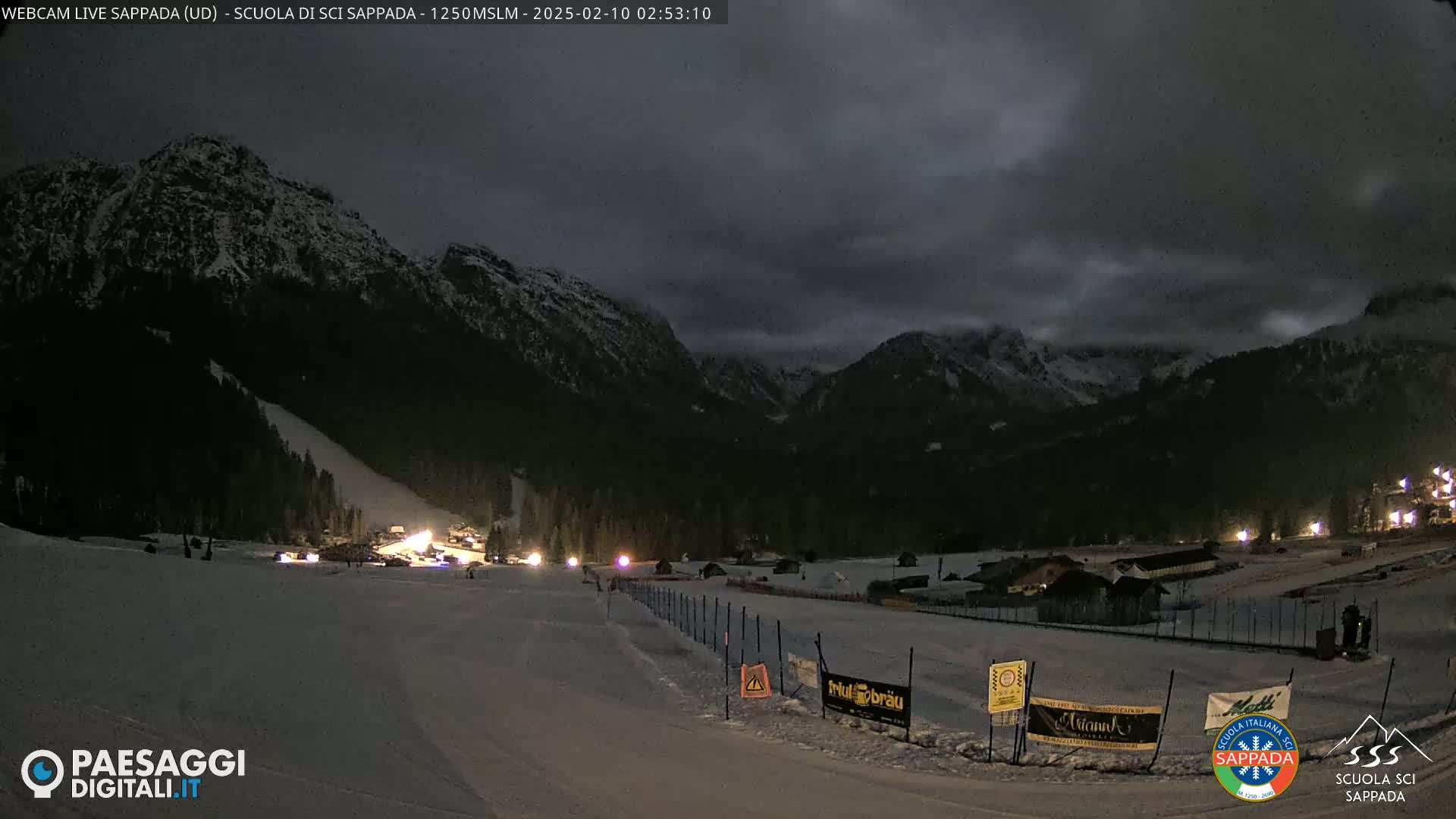 A nighttime view of a snow-covered ski slope nestled in a valley between dark, snow-dusted mountains under a cloudy sky.