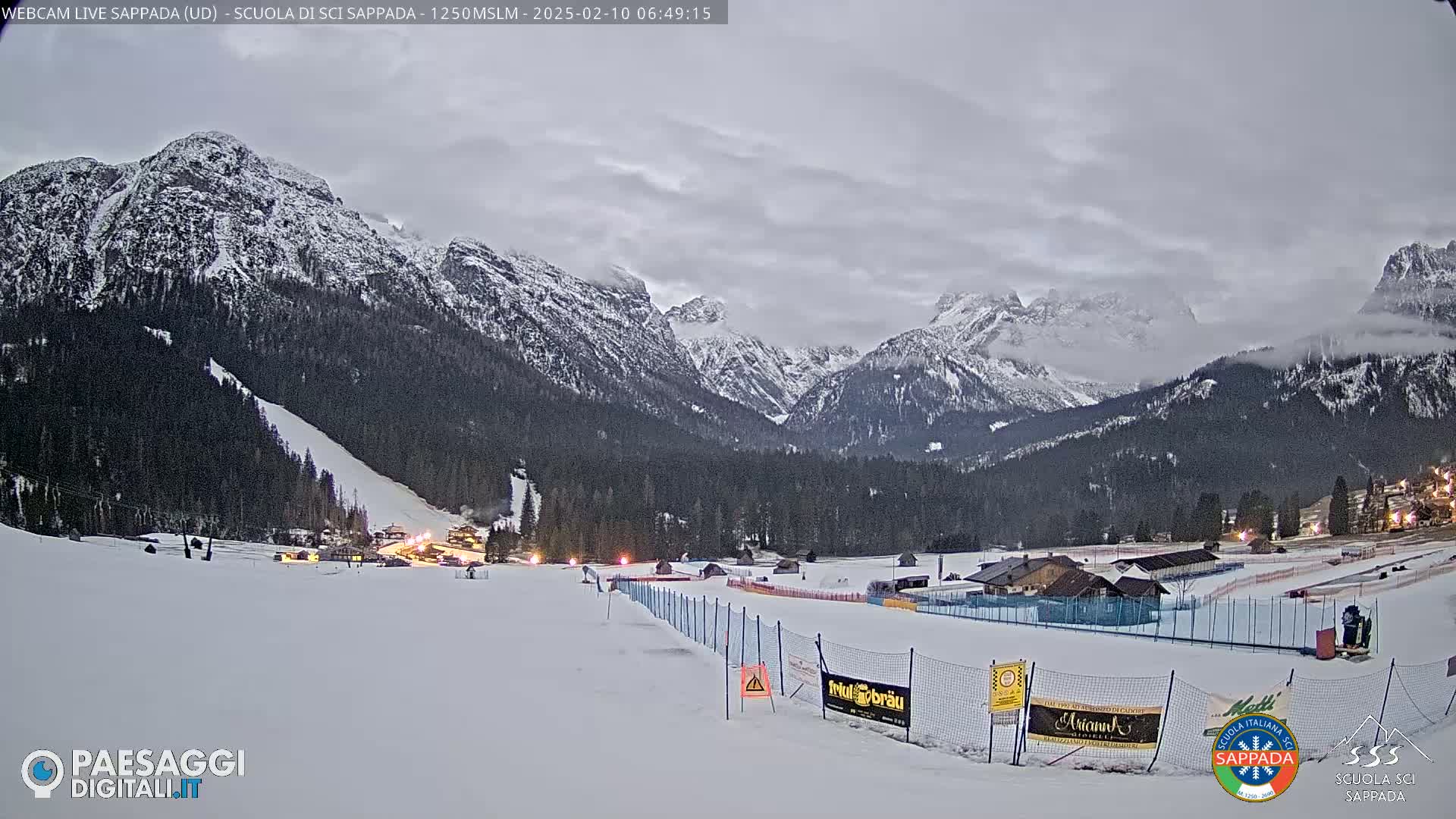 A snow-covered ski slope at the base of snow-capped mountains under a cloudy sky.