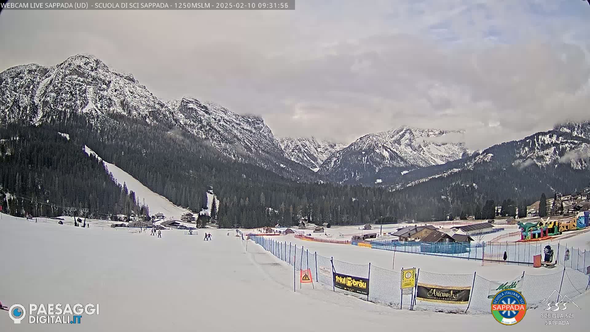 A snowy ski slope and resort nestled in a valley between snow-covered mountains under a cloudy sky.