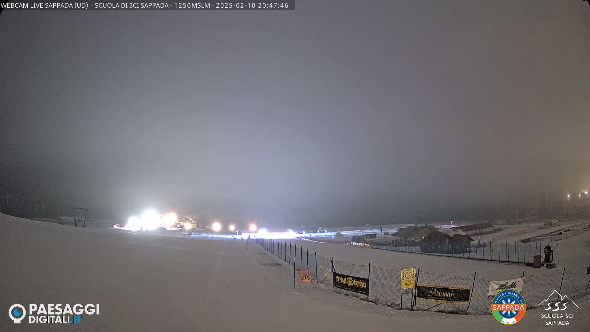 A snowy nighttime scene with fog or mist, showing a dimly lit ski slope with a fence and some buildings in the distance.