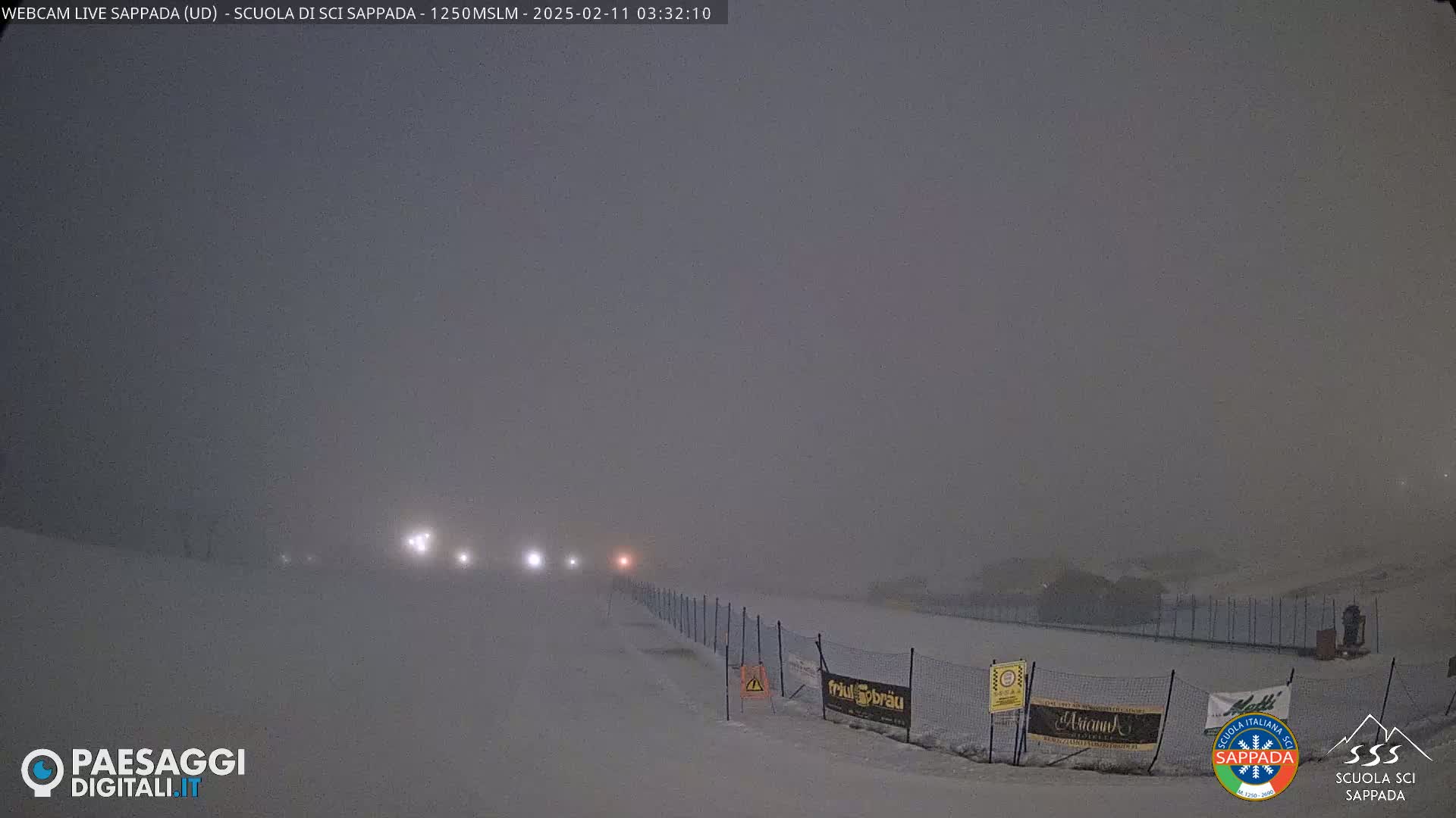 A snow-covered ski slope is dimly lit by distant headlights in heavy snow.