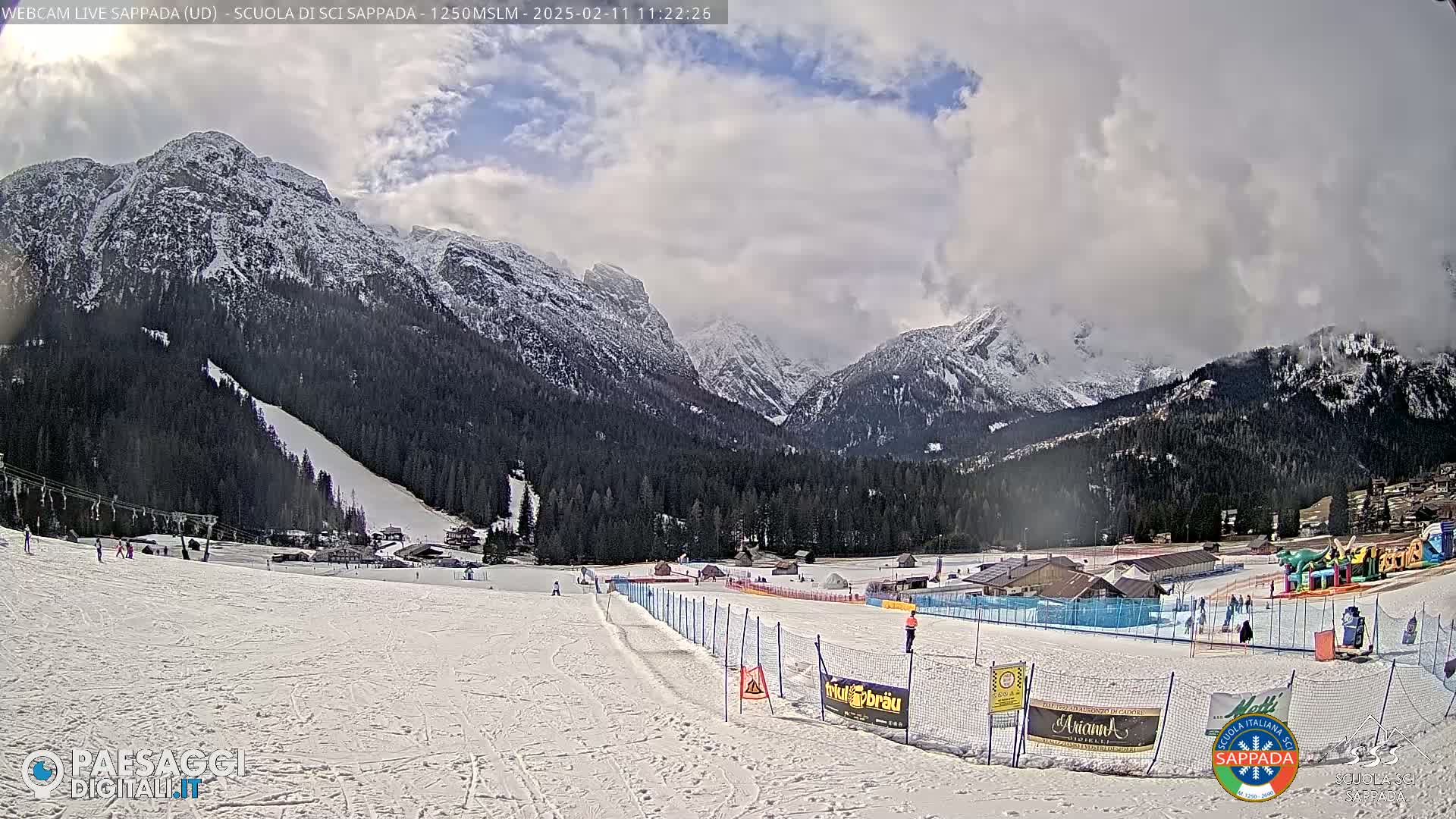 A partly cloudy day shows a snow-covered ski slope in a valley, with snow-capped mountains in the background.