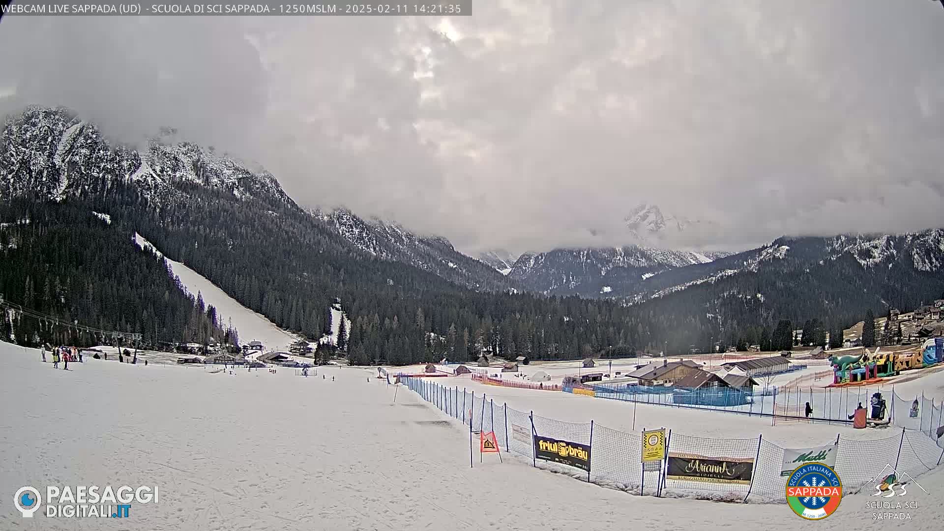 A snowy mountain ski slope with overcast skies shows a ski resort with buildings and some people in the distance.