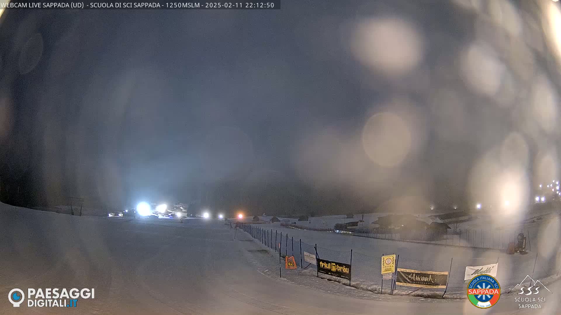 A snowy nighttime scene shows a ski slope with lights illuminating the area under a hazy, dark sky.