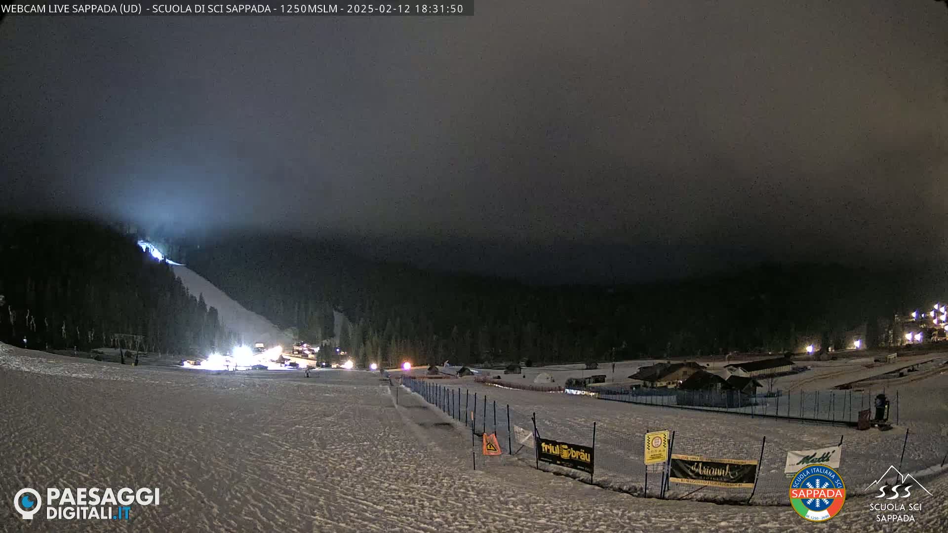 A nighttime view of a snow-covered ski slope illuminated by lights, under a dark, foggy sky.