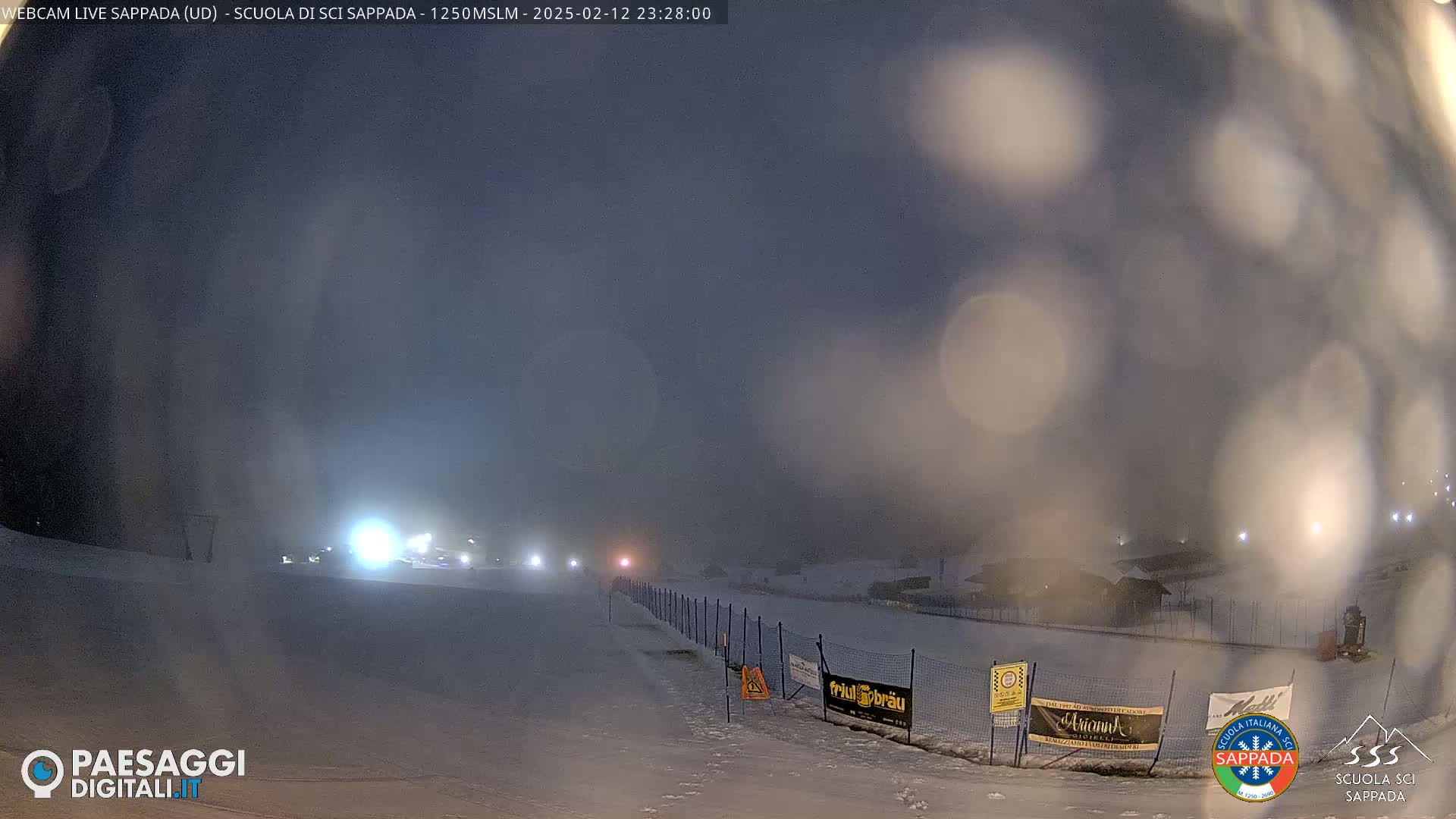 A snowy, nighttime scene shows a dimly lit ski slope with lights in the distance, under a hazy, dark sky.