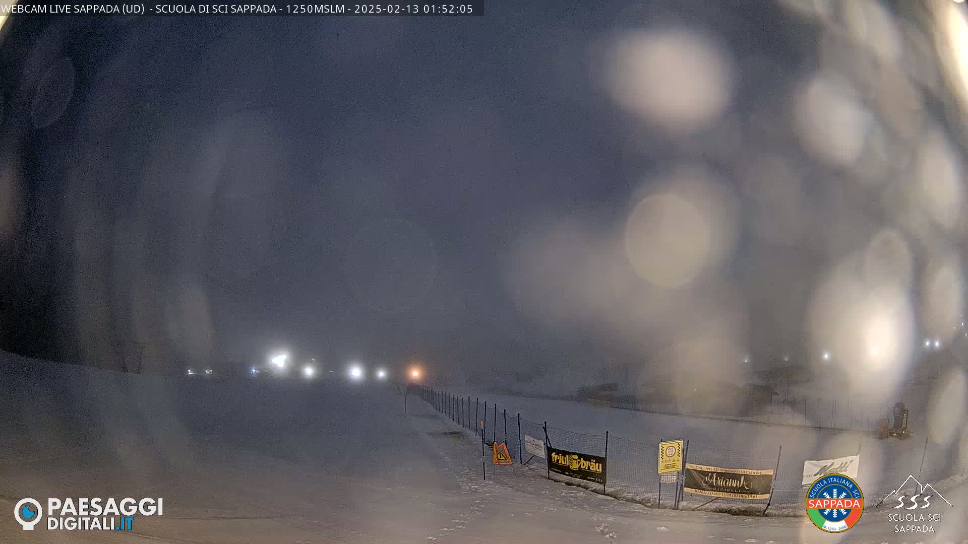 A snow-covered landscape at night is dimly lit by several lights in the distance, with a hazy, snowy atmosphere obscuring visibility.
