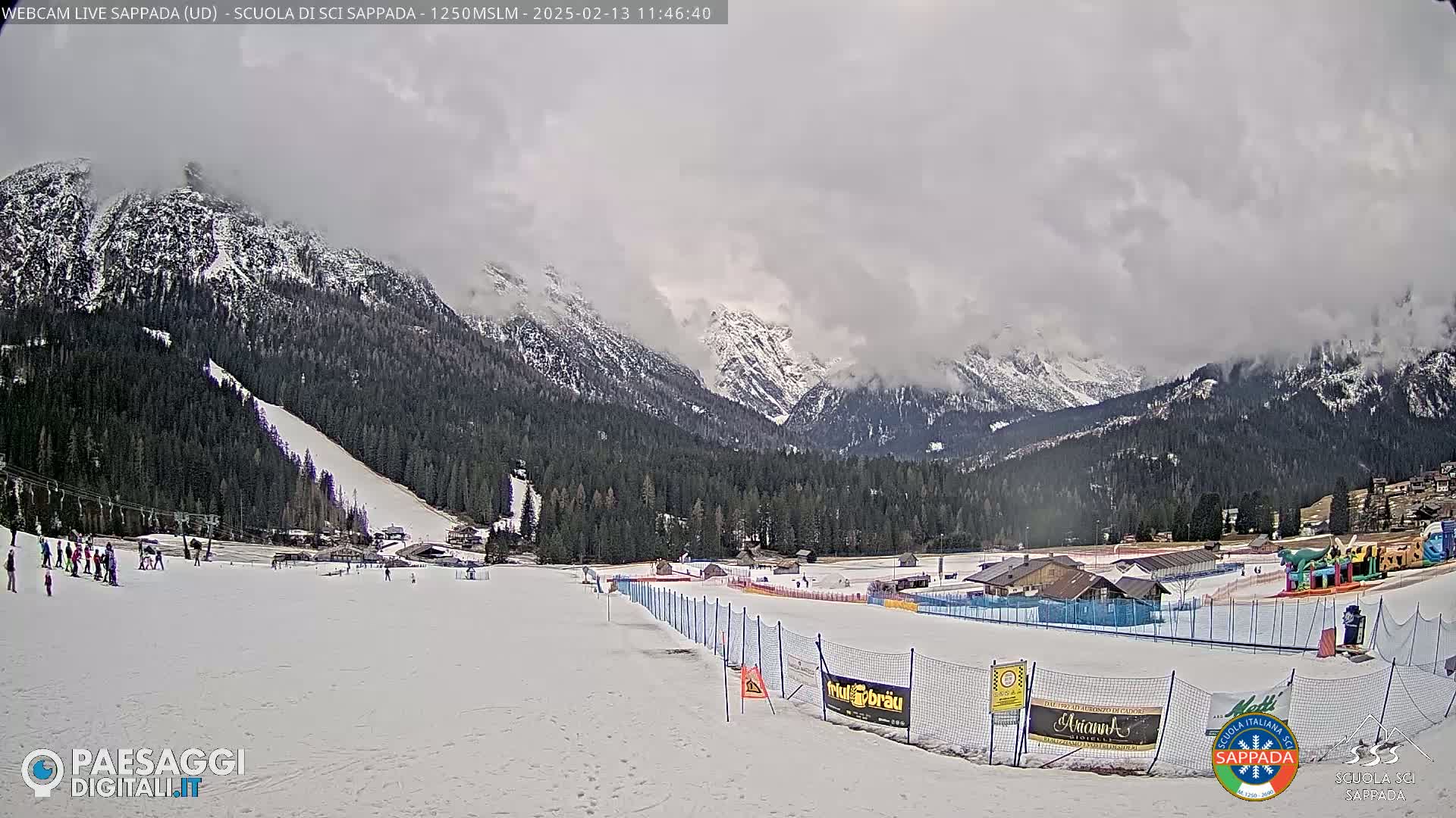 A snow-covered ski slope with a few skiers, nestled in a valley surrounded by snow-capped mountains under an overcast sky.