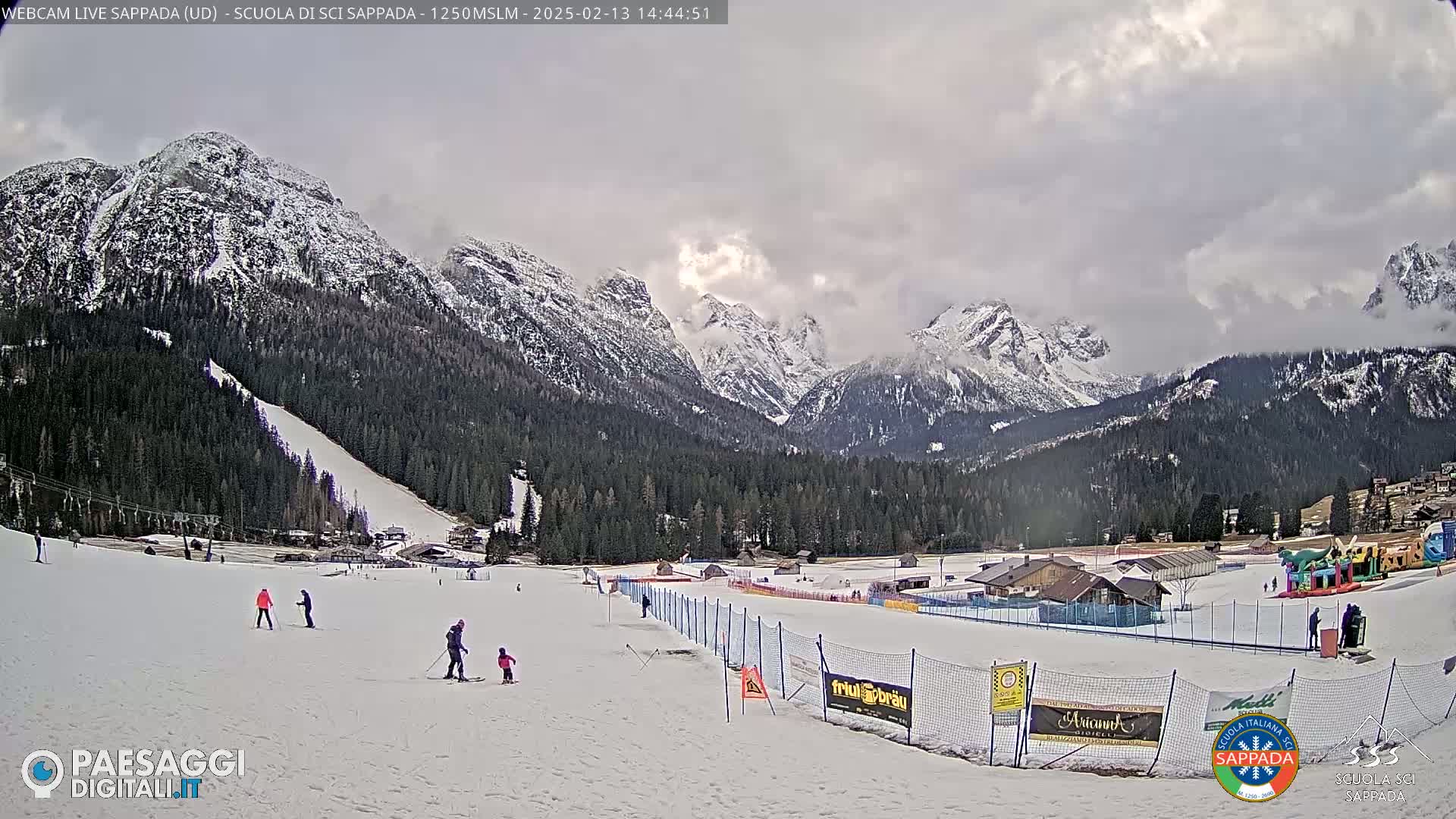 A snowy ski slope with several skiers, a snow-covered mountain range in the background under a cloudy sky.
