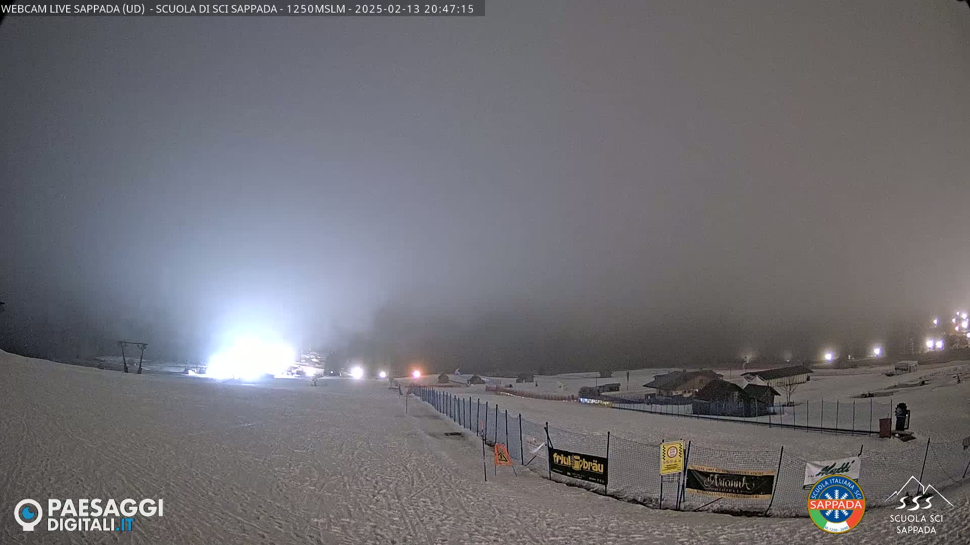 A snowy, foggy nighttime scene at a ski resort shows a slope with lights and fences, and buildings in the distance.