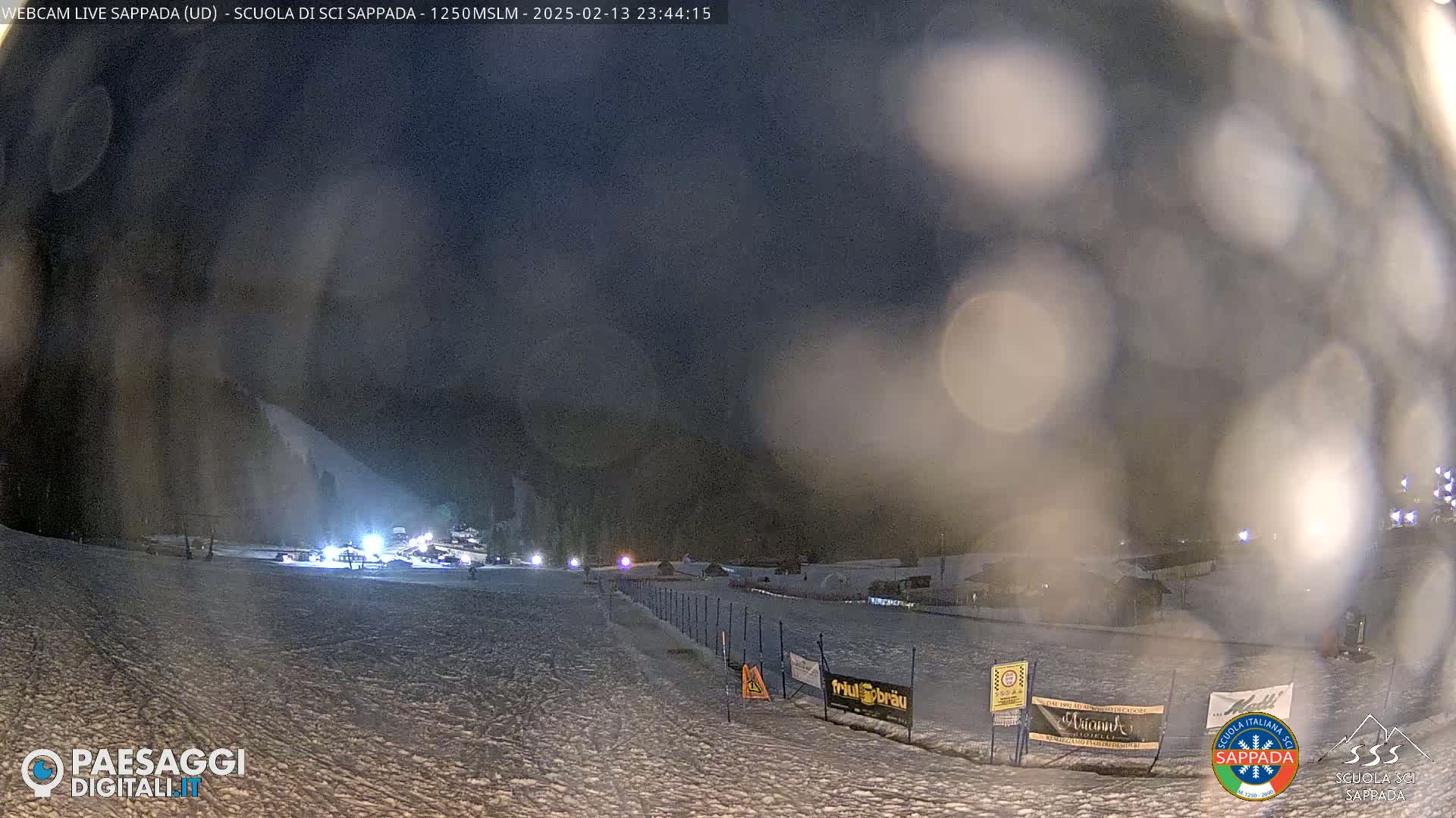 A nighttime view of a snow-covered ski slope with lights illuminating the base area, under hazy, possibly snowy conditions.