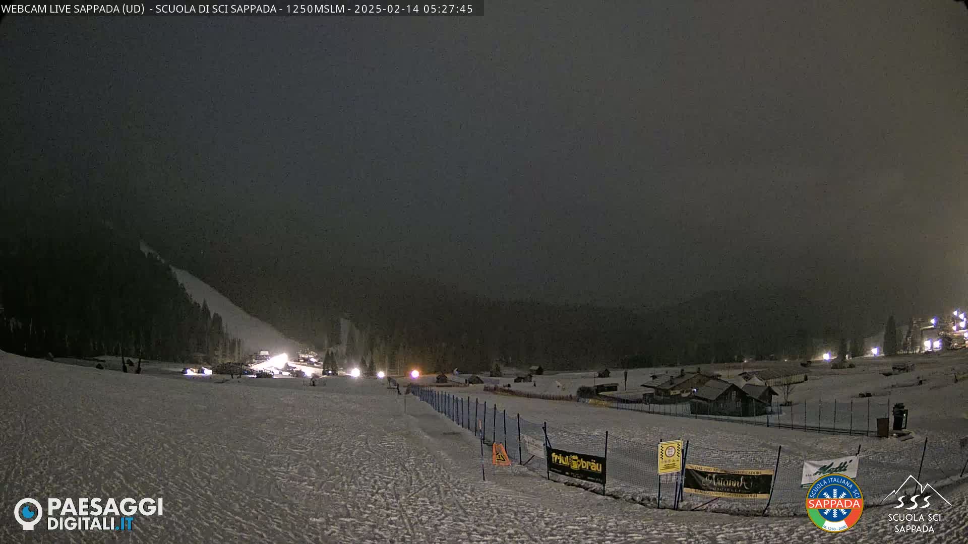 A snow-covered ski slope at night under a dark, overcast sky, with lights illuminating a few buildings and vehicles in the distance.