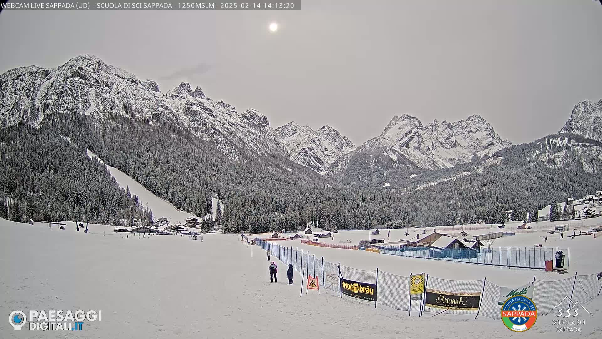 A snow-covered ski slope with a few people, surrounded by snow-capped mountains under a mostly cloudy sky.