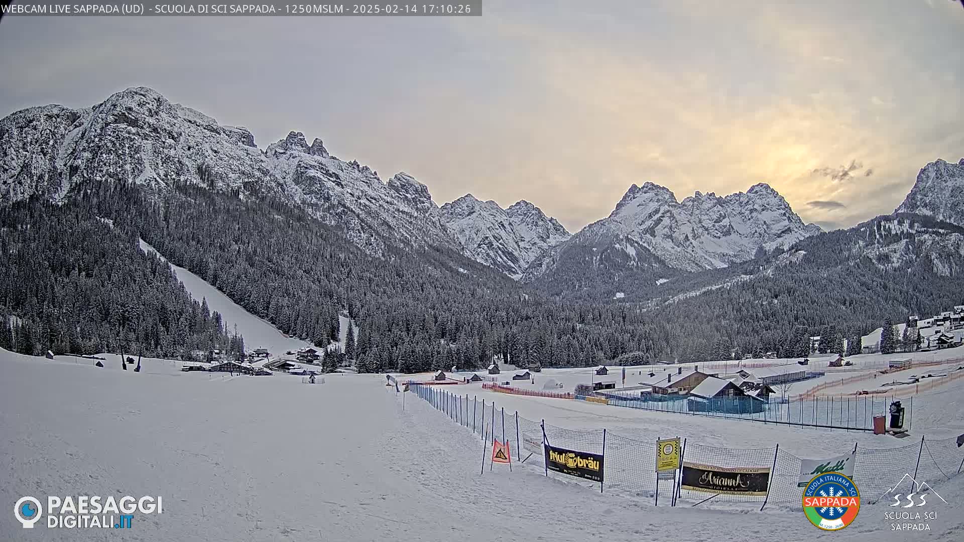 A snow-covered ski slope and surrounding snow-capped mountains are visible under a partly cloudy, sunset sky.
