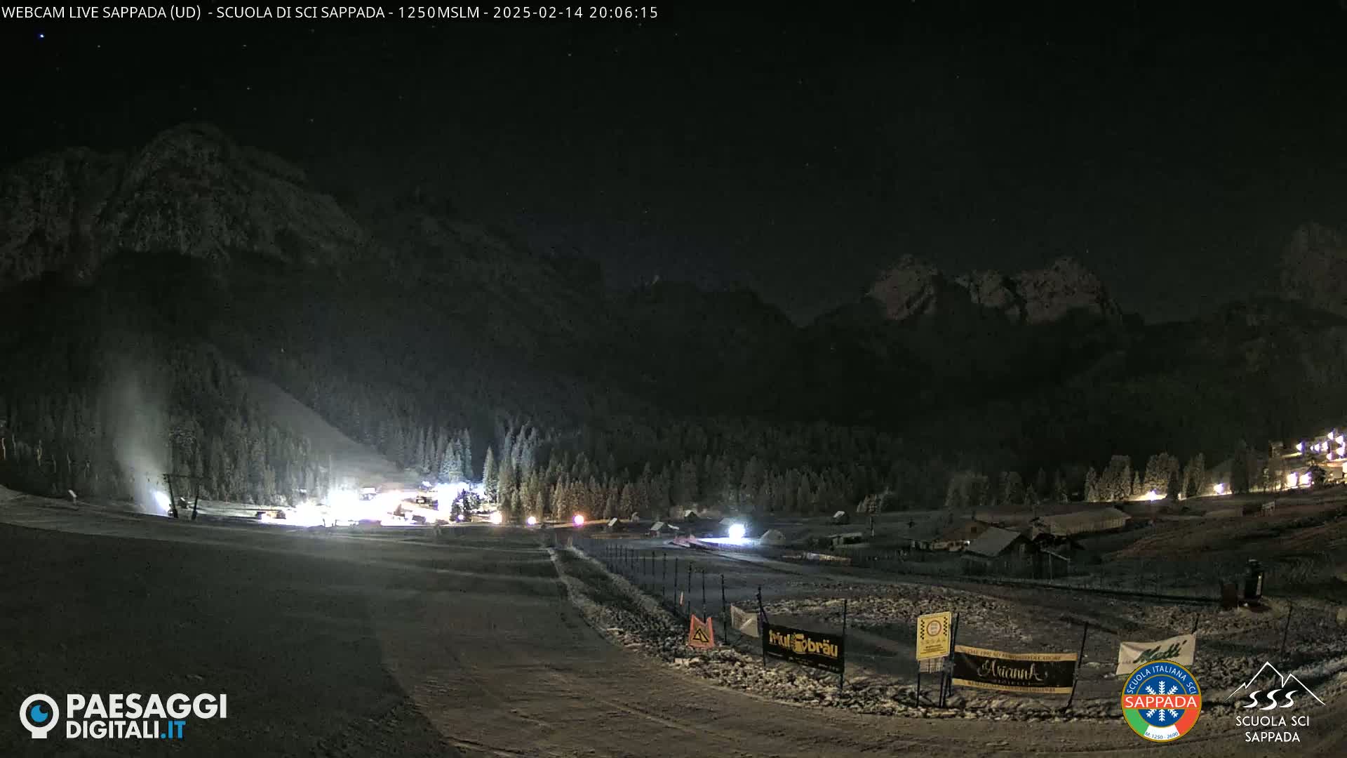 A snow-covered ski slope at night is illuminated, with mountains in the background under a clear, dark sky.
