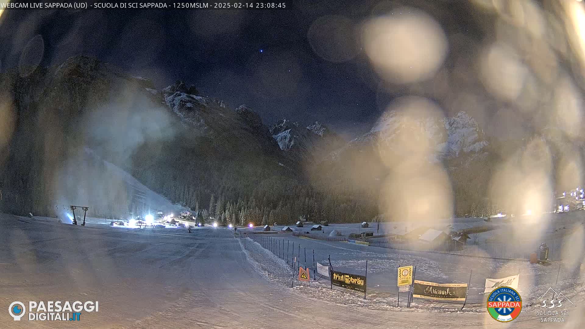 A snow-covered ski slope at night under a starry sky, with mountains in the background and some lights visible in the distance.