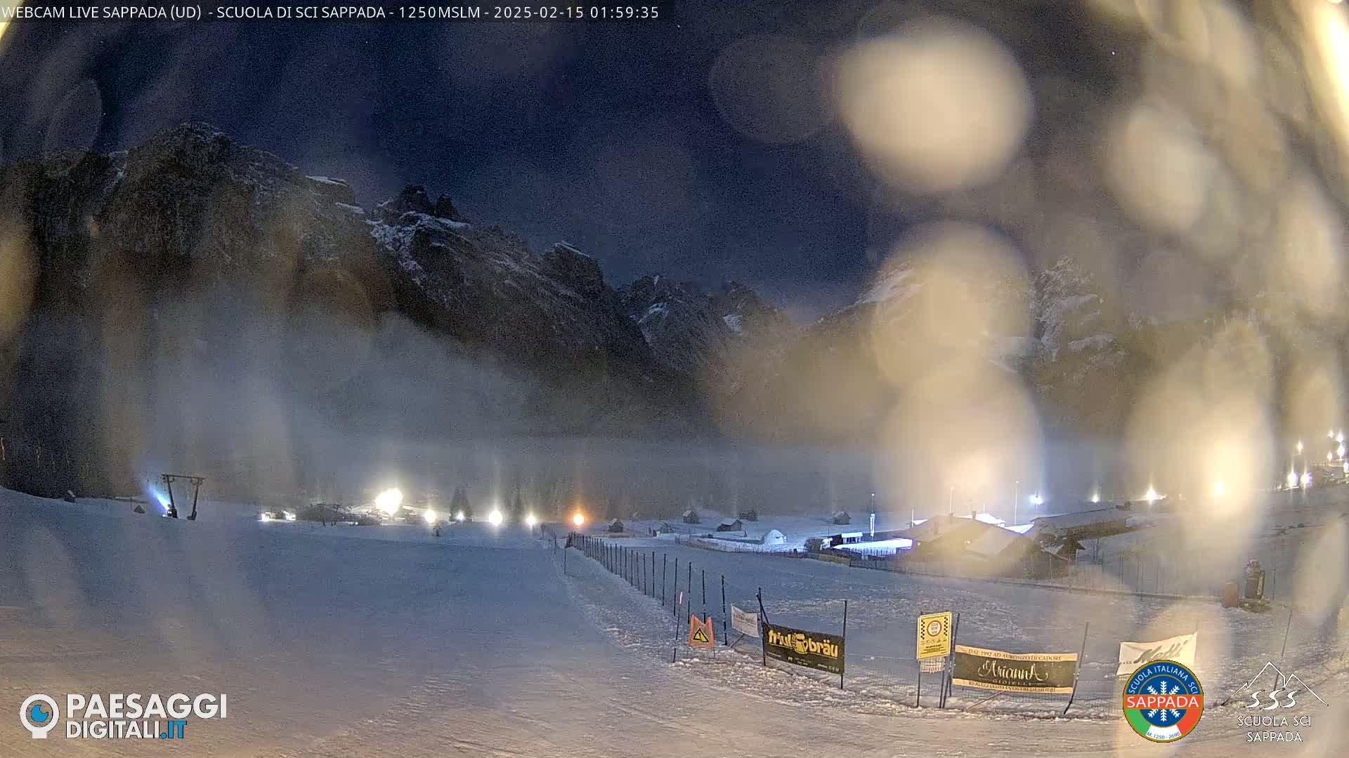 A snow-covered ski slope at night is illuminated by lights, with snow falling and mountains in the background under a dark sky.