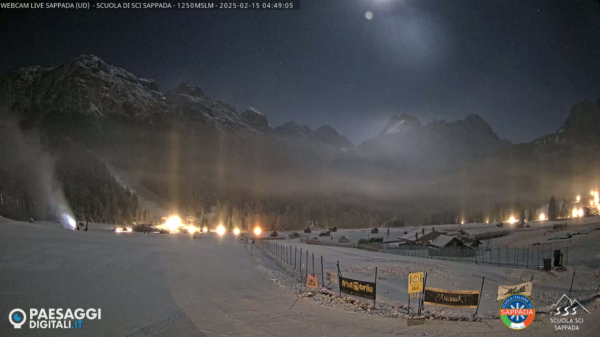 Snow-covered hills and mountains are illuminated by lights at night under a bright moon, with snowmaking machines operating on a ski slope.