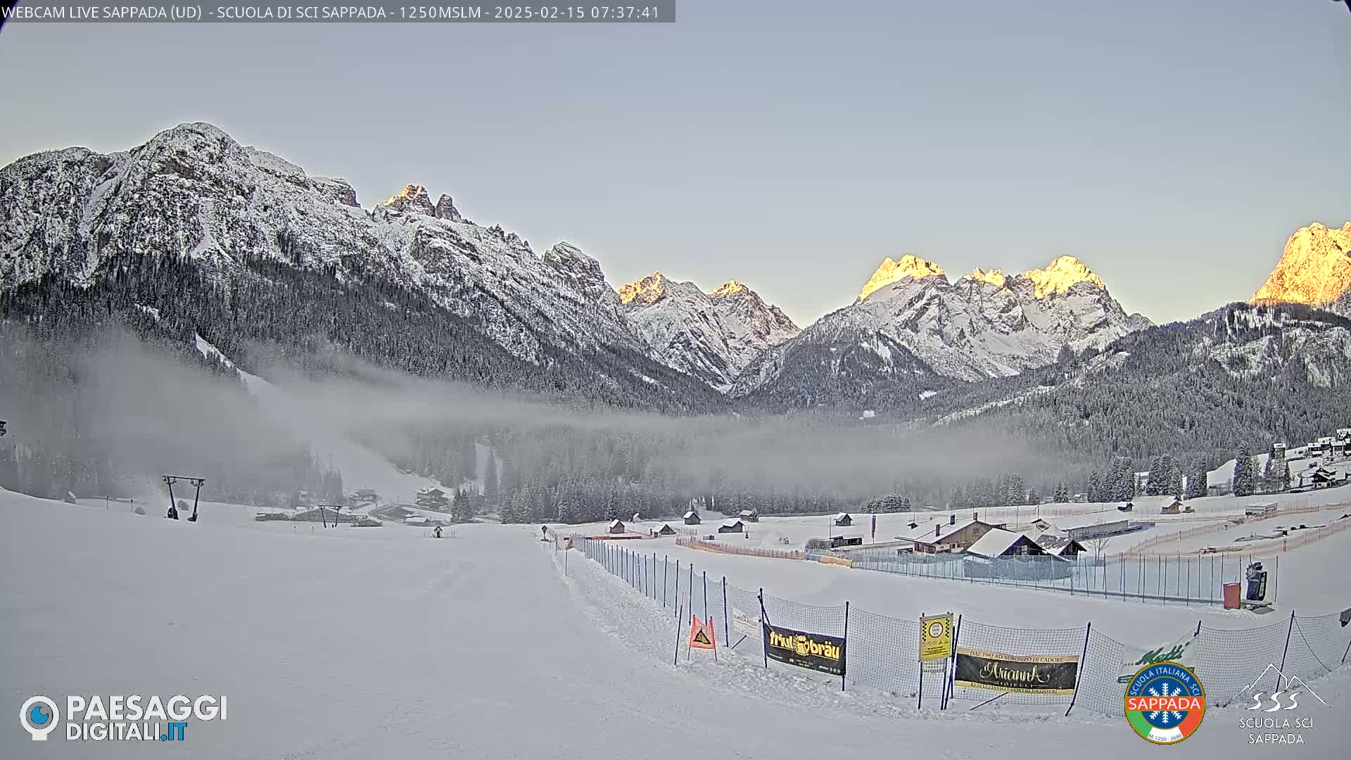 A snow-covered ski slope with a low-lying fog sits in front of sunlit, snow-capped mountains under a clear, pale-blue sky.