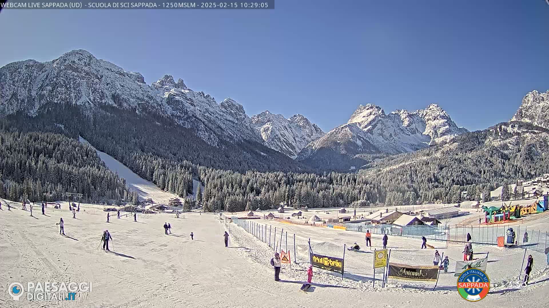 A snow-covered ski slope with skiers and snowboarders, surrounded by snow-capped mountains and evergreen trees under a clear blue sky.