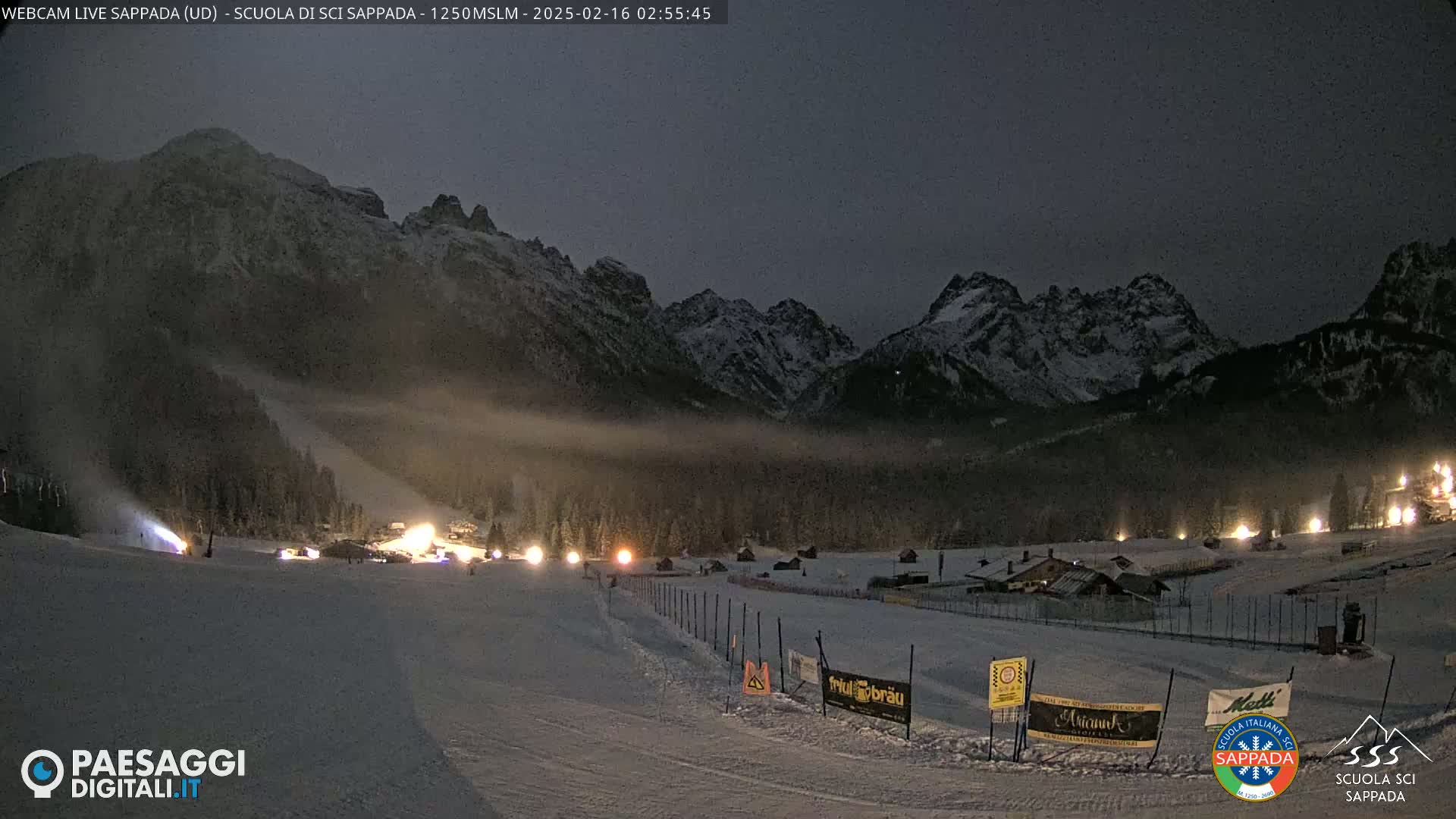 A snow-covered ski slope at night, illuminated by lights, with snow-capped mountains in the background under a dark sky.