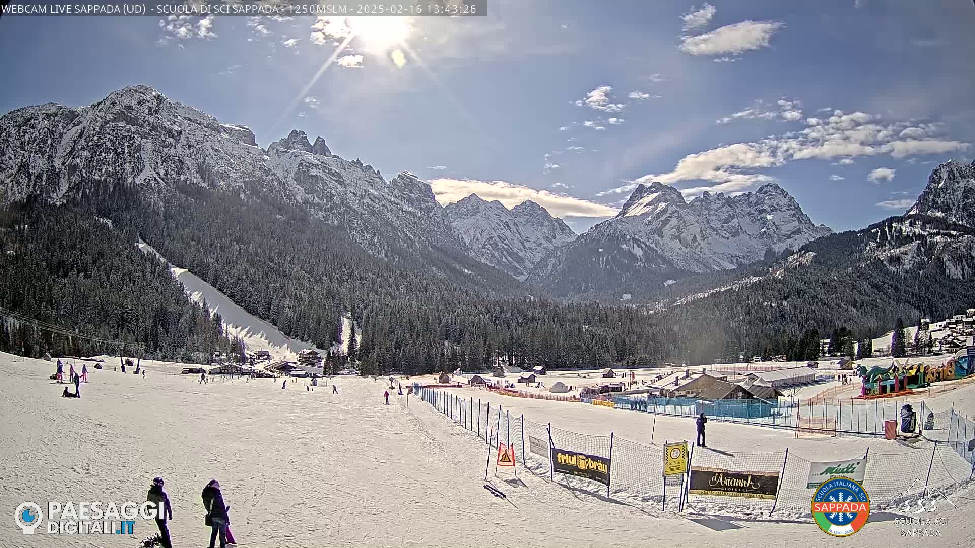 A sunny day in a snow-covered mountain valley shows a ski slope with several people scattered across it, a ski resort in the mid-ground, and snow-capped mountains in the background.