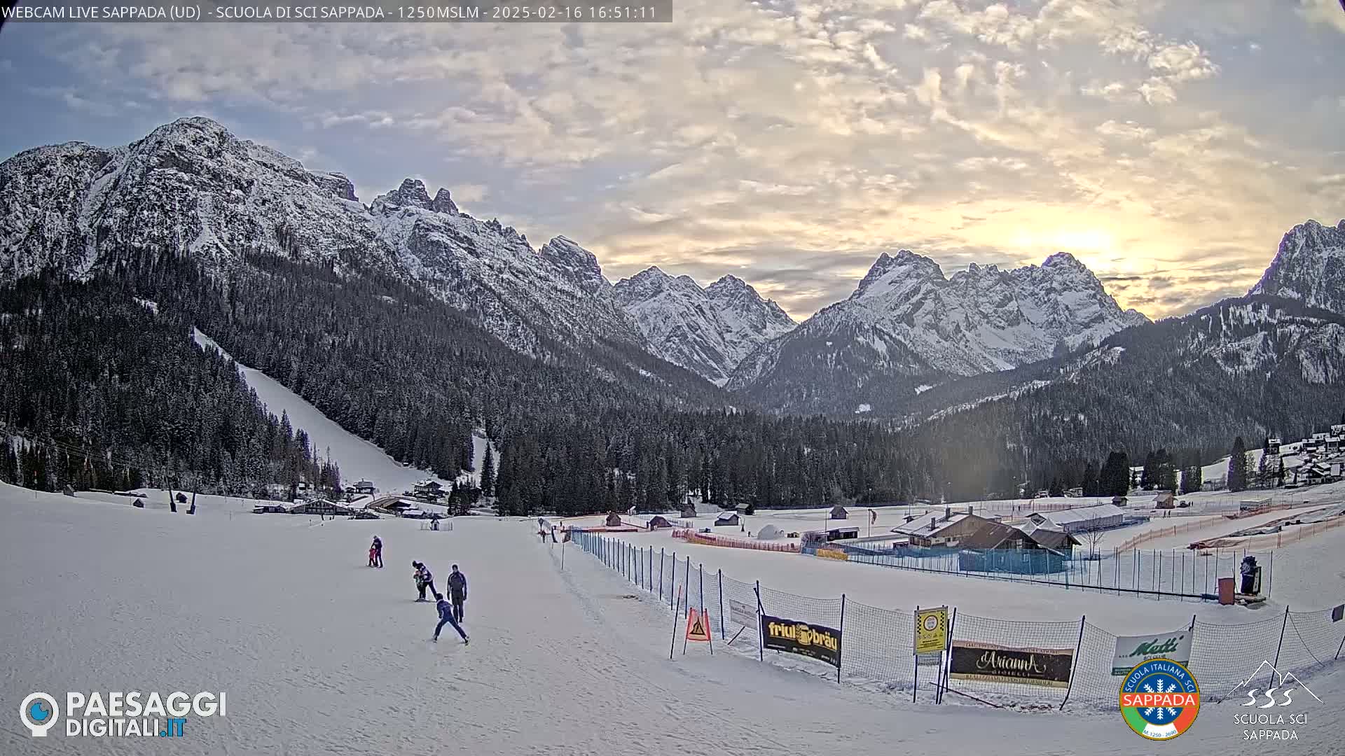 A snow-covered ski slope with several skiers, a few buildings, and a mountain range in the background under a partly cloudy sky at sunset.