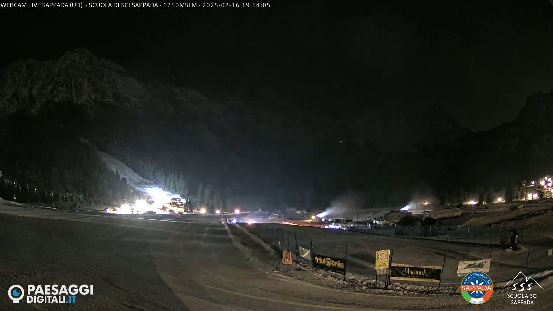 A nighttime view of a snow-covered ski slope with artificial lighting, illuminated buildings, and a snow-covered mountain in the background.