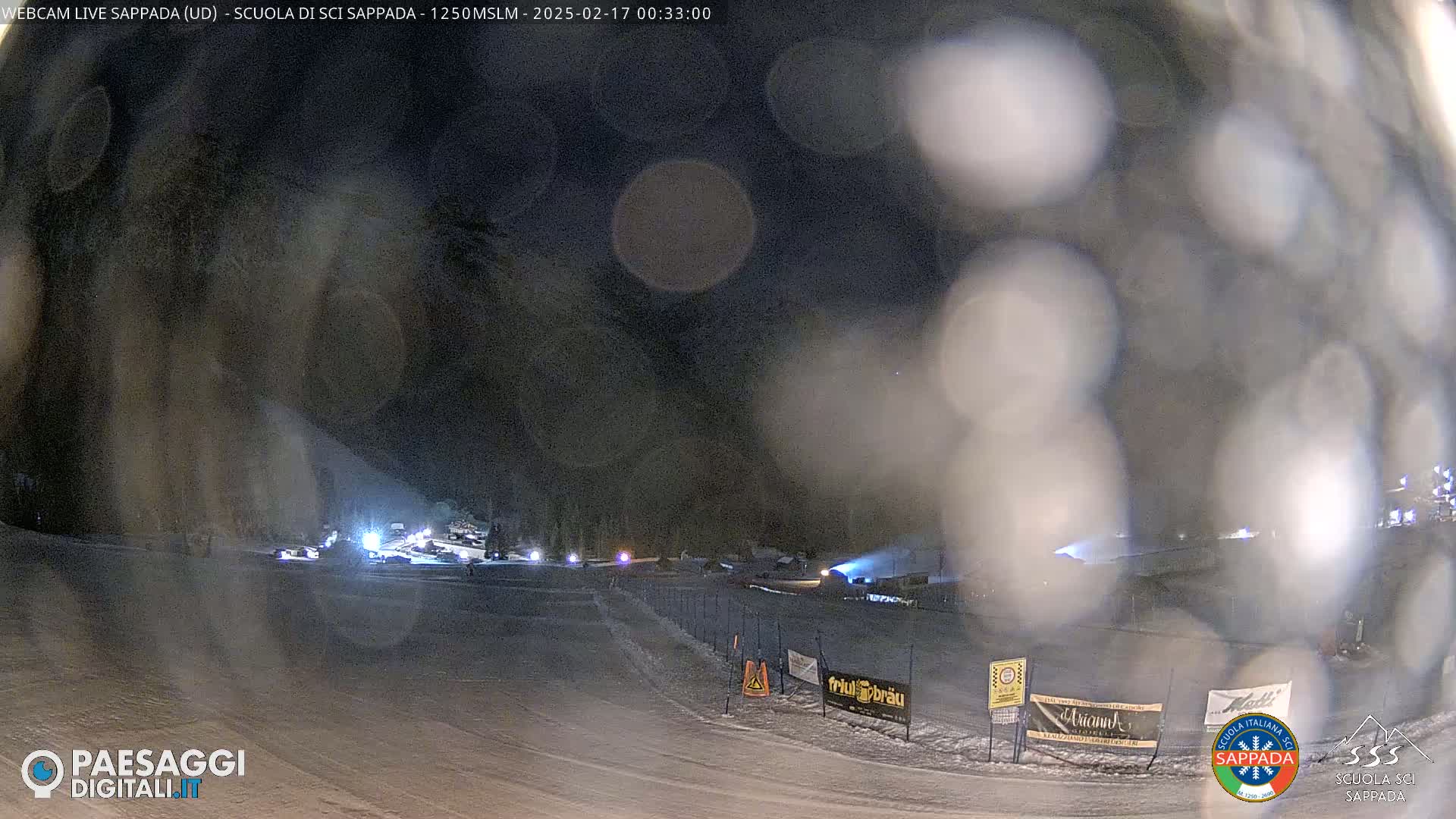A nighttime view of a snow-covered ski area with some lights and blurry snow in the air.