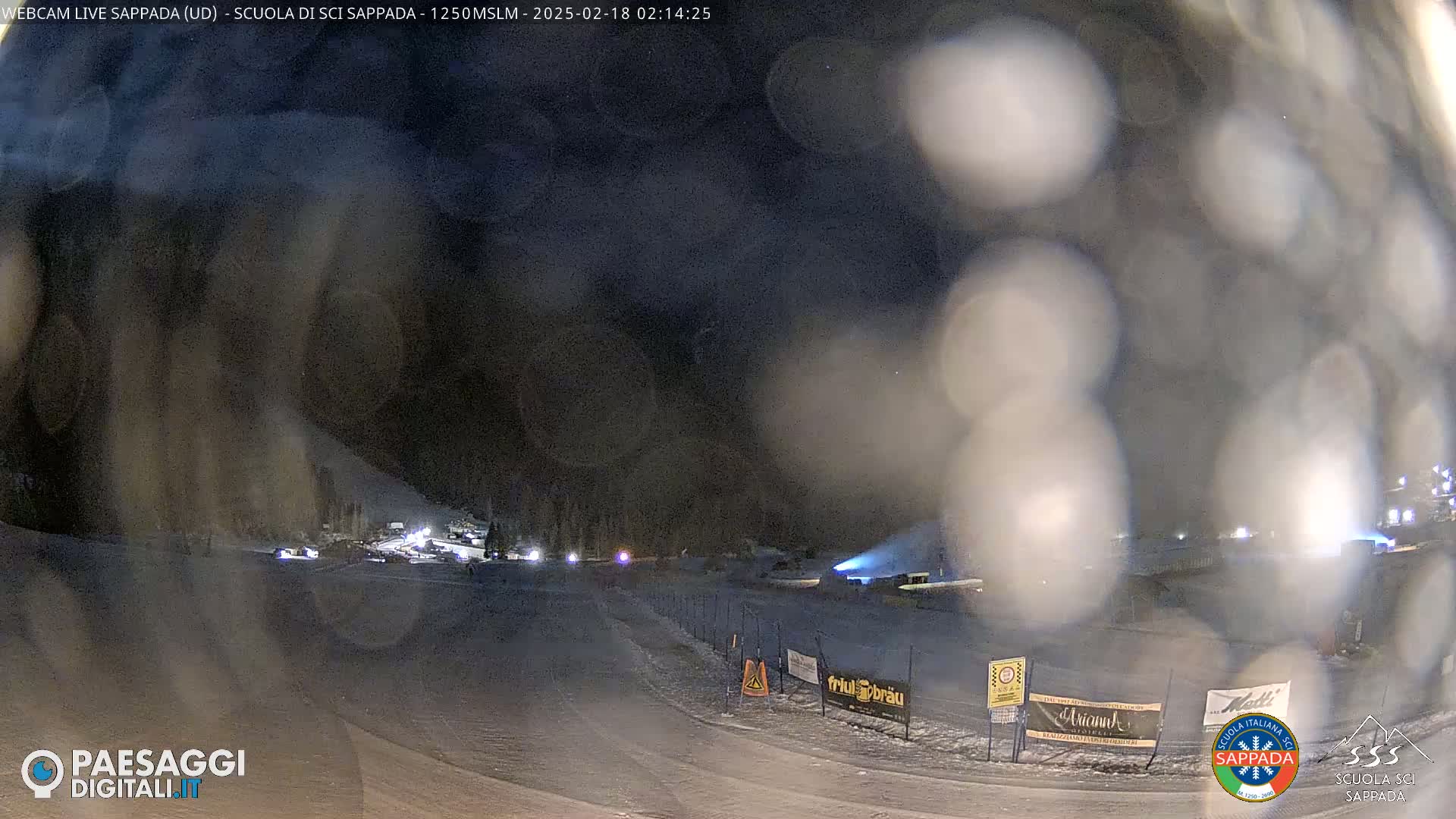 A nighttime, snowy scene shows a road leading to a lit-up area with several buildings, partially obscured by blowing snow or precipitation.