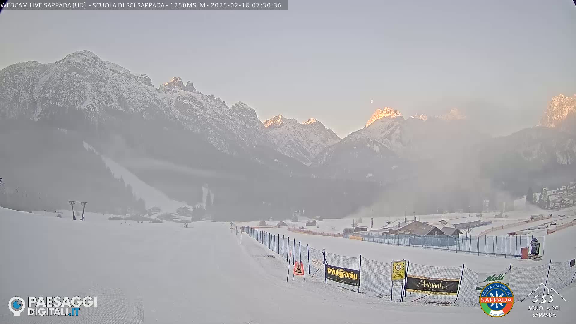 A snow-covered ski slope with a hazy sunrise illuminating snow-capped mountains in the background.