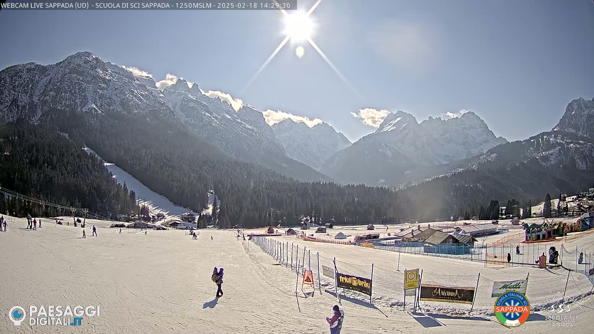 A sunny day at a snow-covered ski resort in the mountains, with skiers and snowboarders visible on the slopes.