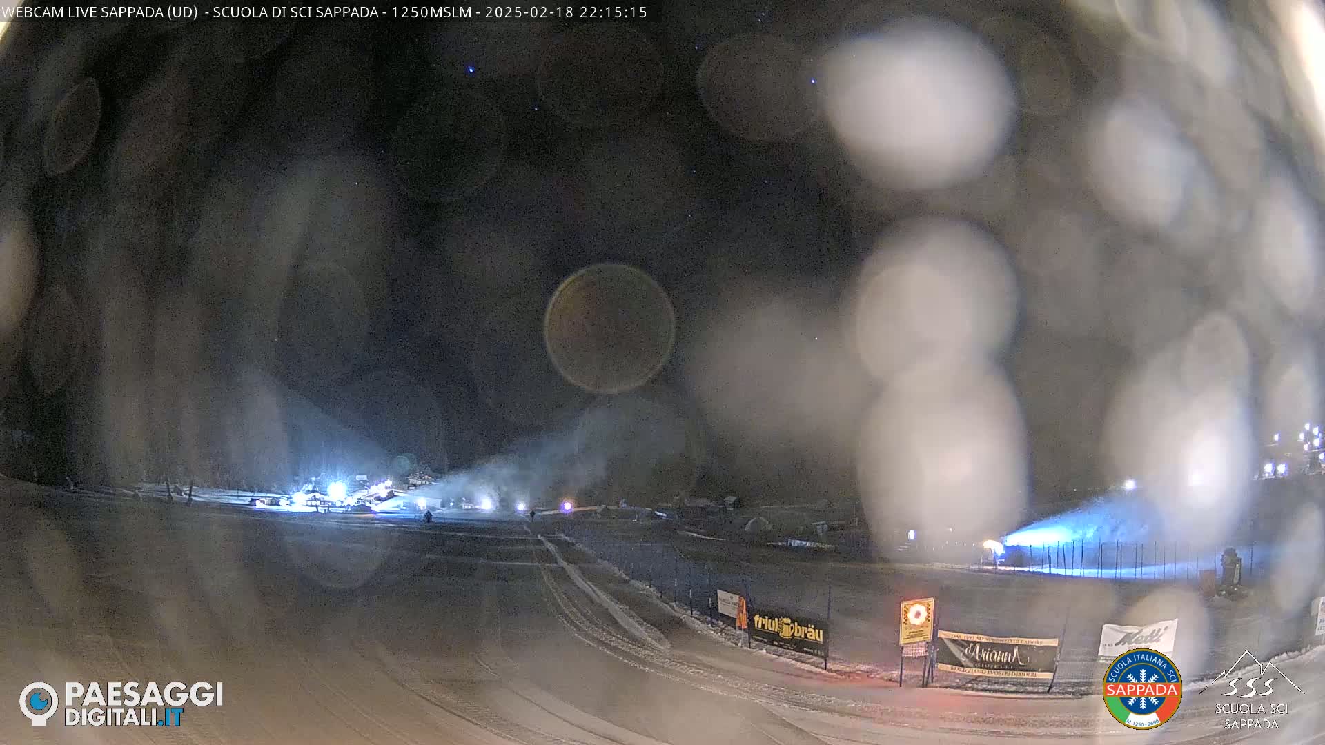A snow-covered nighttime scene shows snowmaking machines operating on a ski slope under a partly cloudy sky.