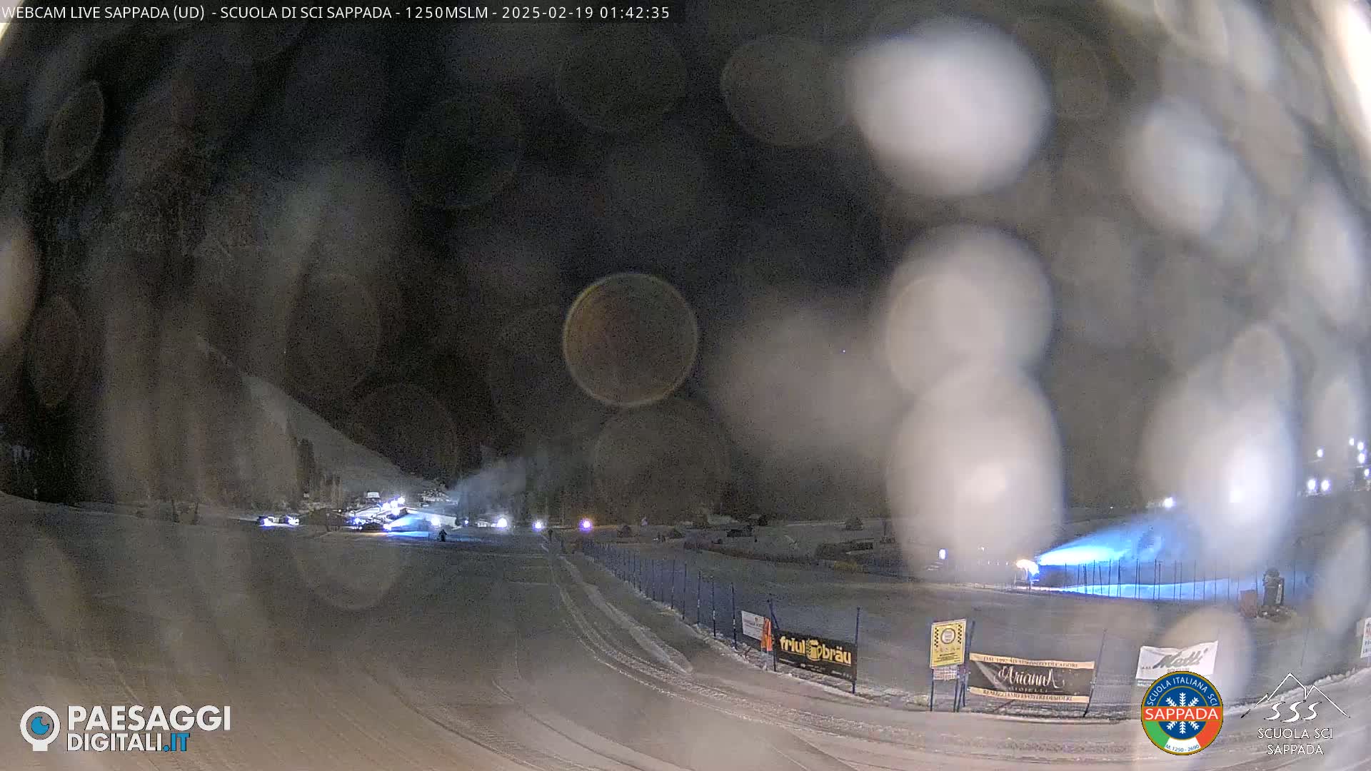 A nighttime view of a snow-covered ski slope with snowmaking machines operating under hazy conditions.