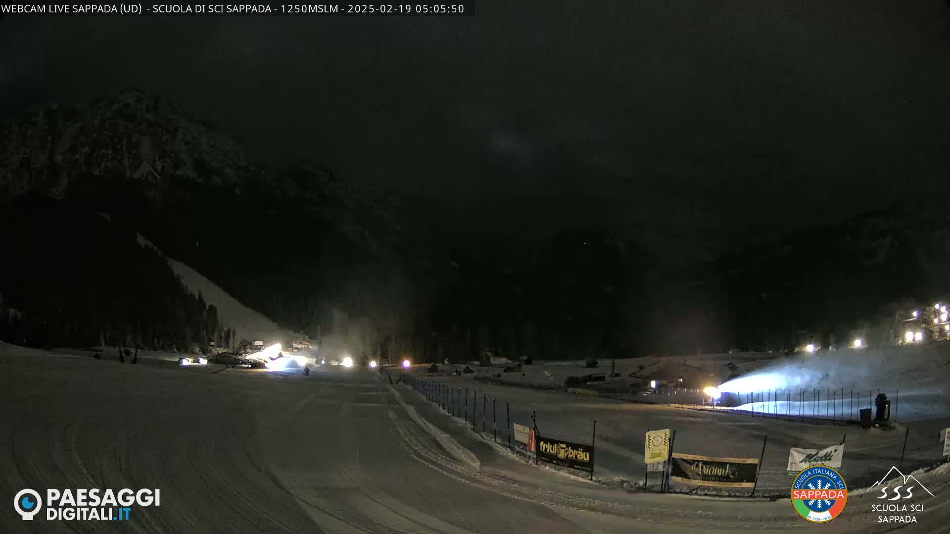 A nighttime view of a snow-covered ski slope with snowmaking machines operating under a dark, clear sky.